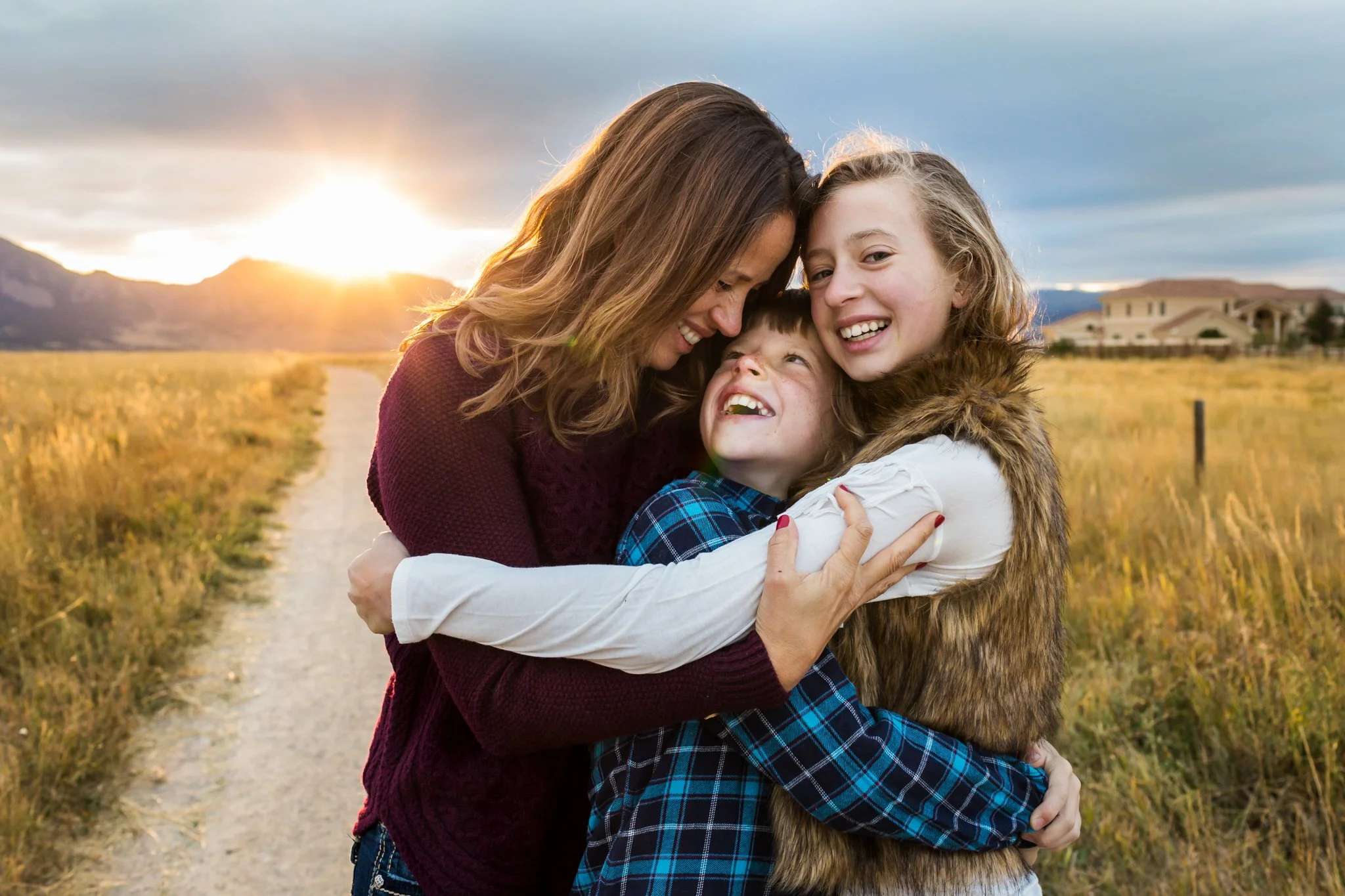 A woman and two girls hugging outdoors in a field during sunset, smiling and showing affection.