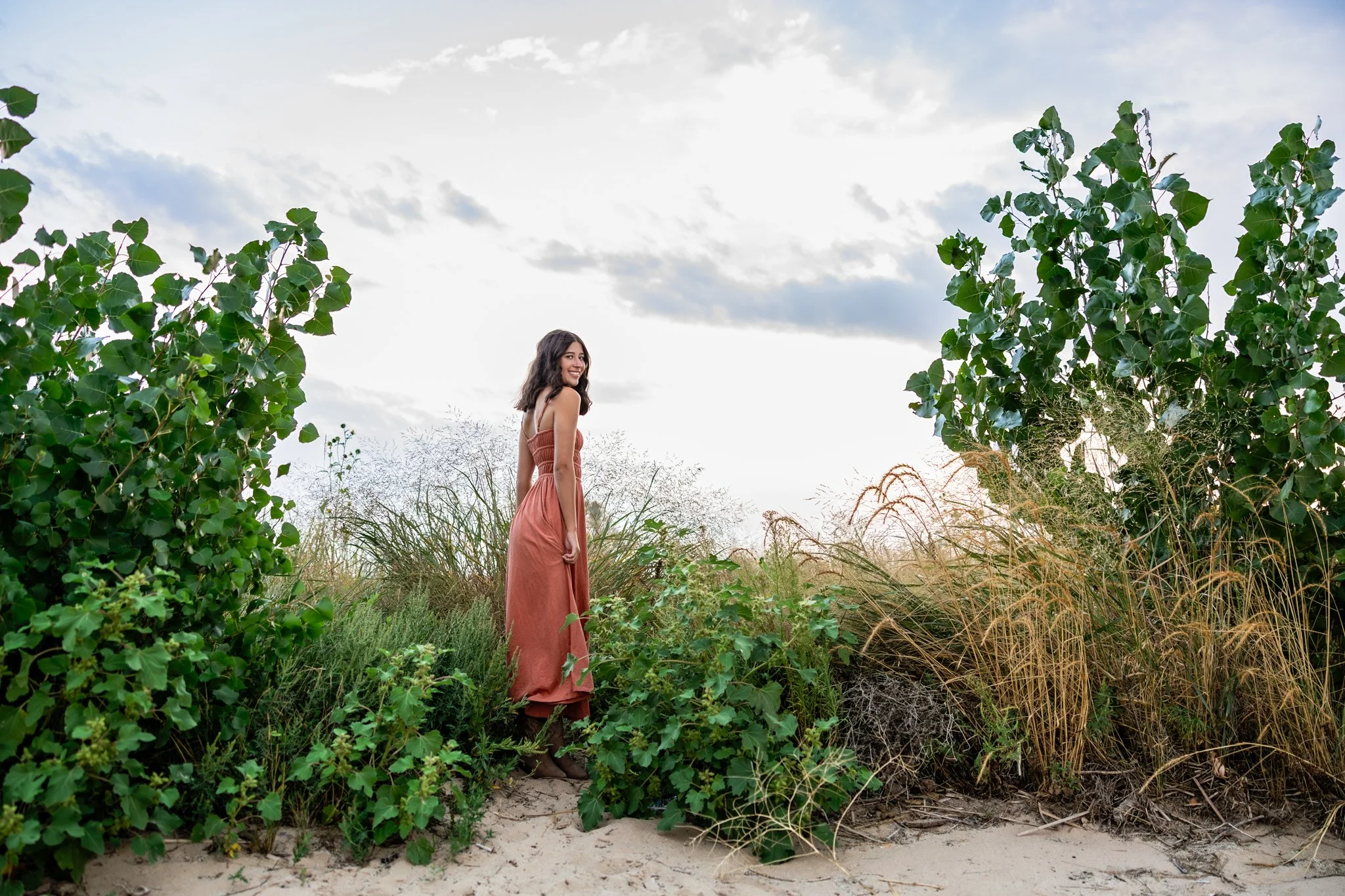 Denver Senior Photographer captures portrait of senior girl in muted red dress standing among bushes.