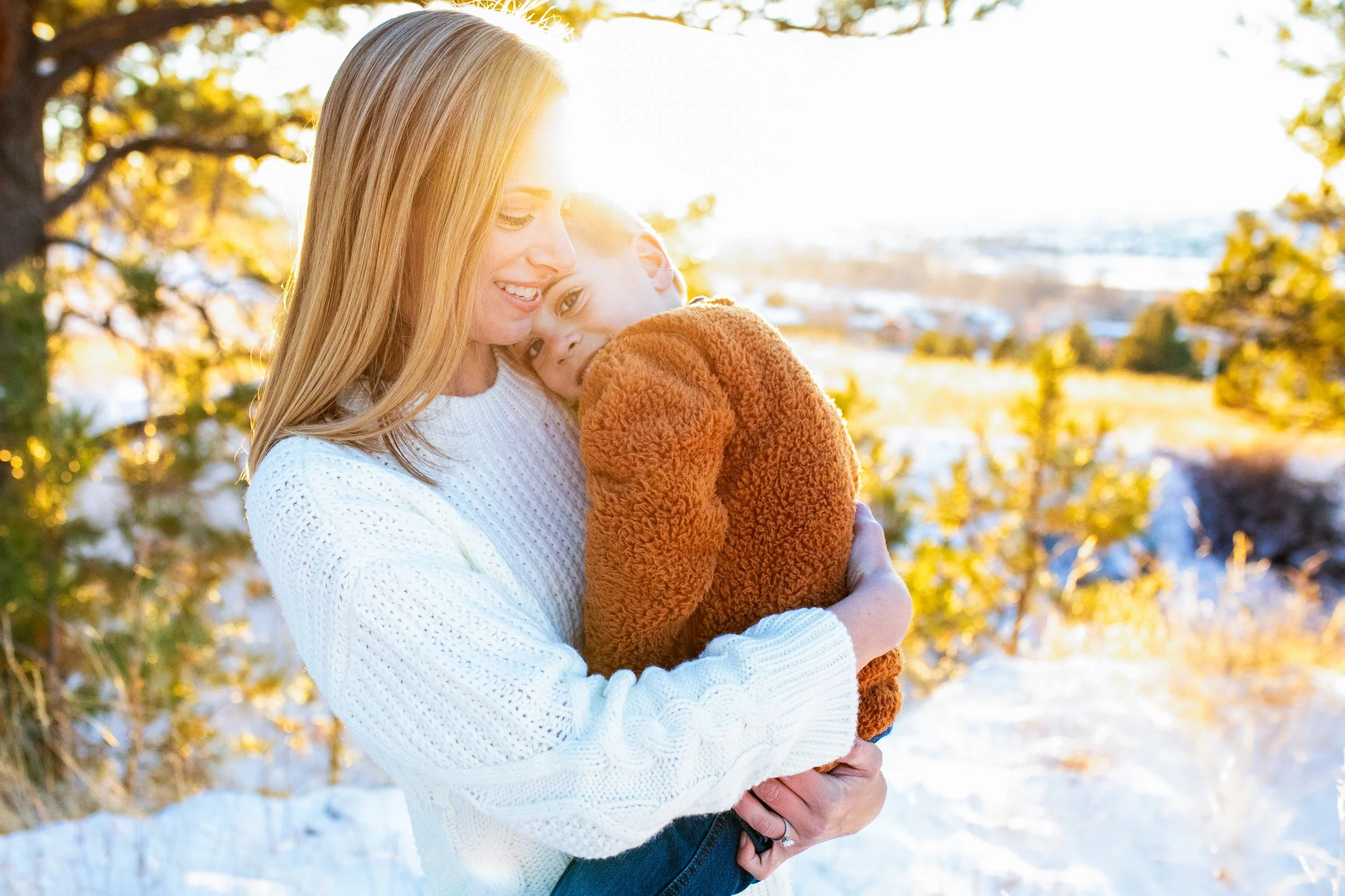 A woman with blonde hair, wearing a white sweater, holding a young boy with brown hair and a brown fuzzy jacket, in an outdoor setting with trees and snow, during sunset.