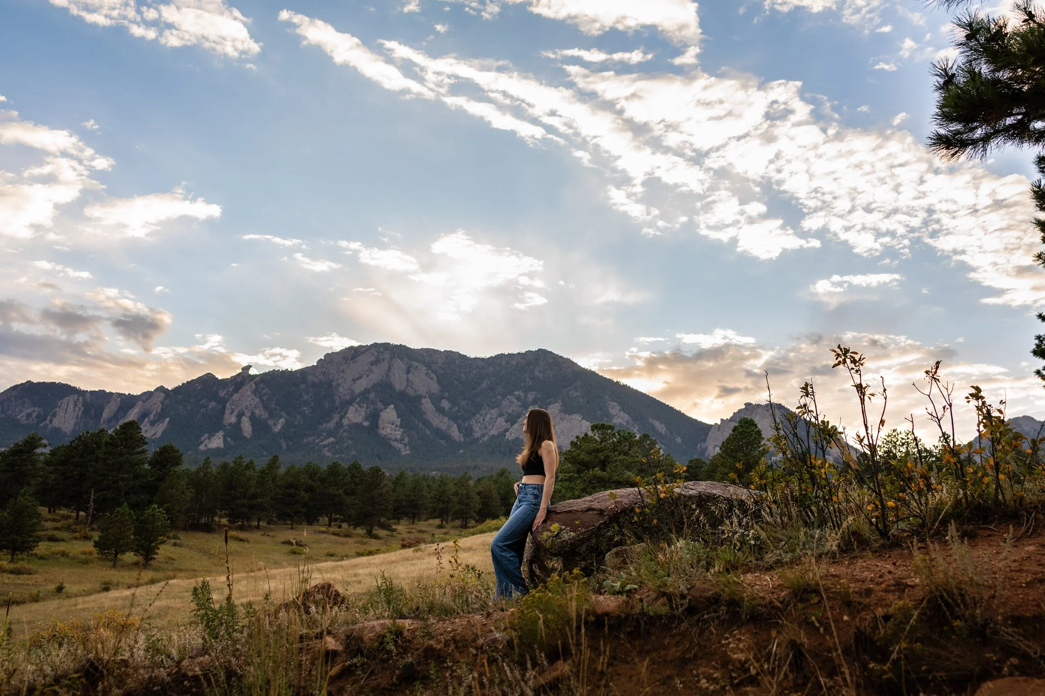 Colorado Senior Photographer takes photograph of senior girl leaning up against a rock in a field at sunset with sweeping views of the mountains behind her.
