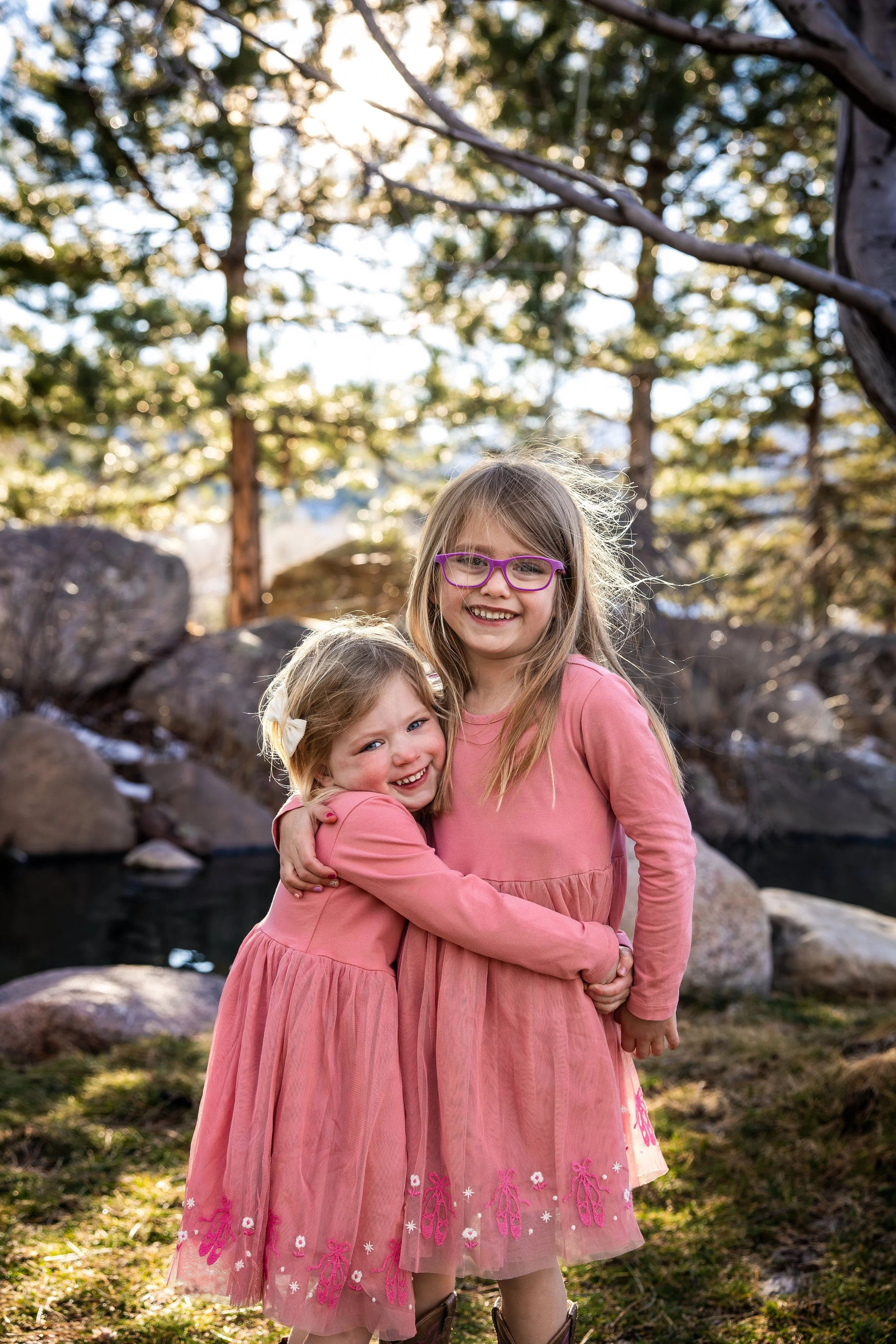 Two young girls wearing matching pink dresses hugging each other outdoors near trees and rocks, smiling at the camera.