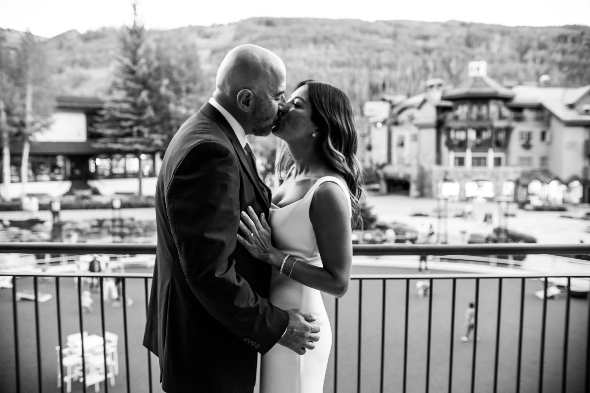 A black and white photo of a couple kissing on a balcony with a scenic background of buildings, trees, and mountains.