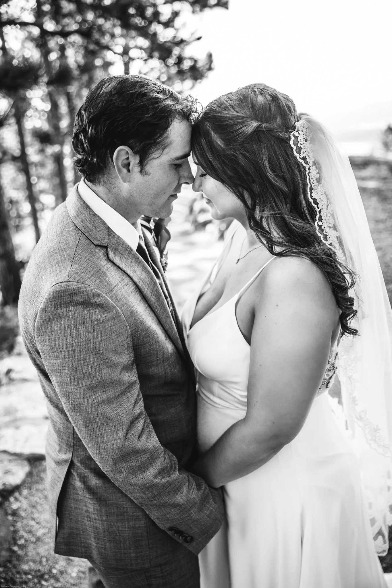 A black and white photo of a bride and groom touching foreheads with eyes closed, outdoors with trees in the background.