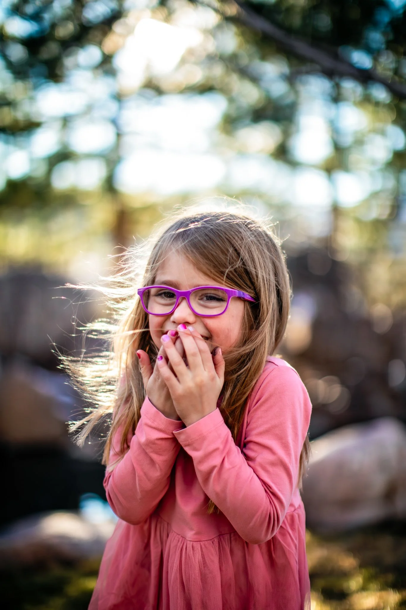 A young girl with glasses and long hair, wearing a pink dress, laughs with her hand covering her mouth outdoors with blurred trees and sunlight in the background.