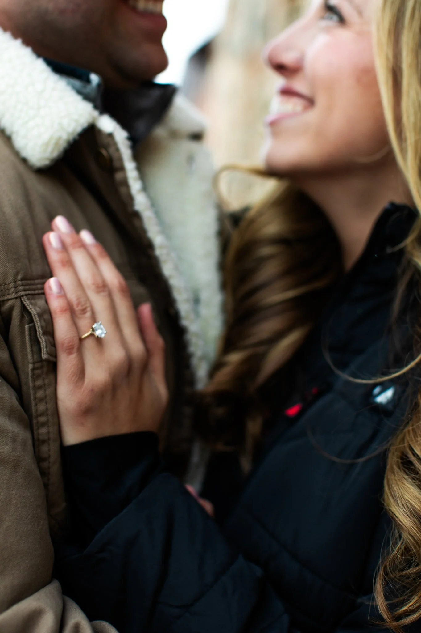 A close-up of a couple smiling at each other, with the woman showing her engagement ring.