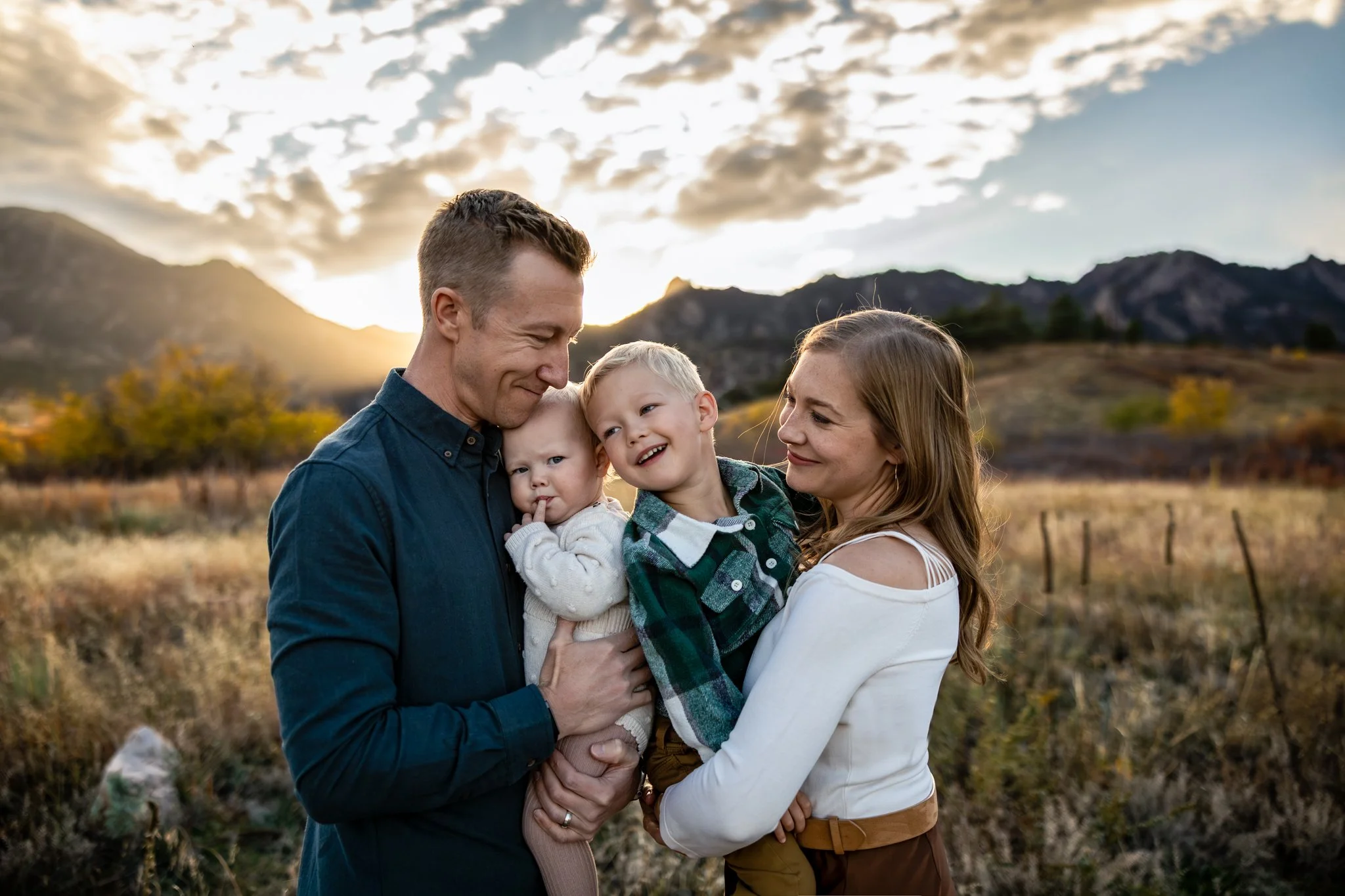 A family of four smiling and embracing outdoors during sunset, with mountains and a partly cloudy sky in the background.