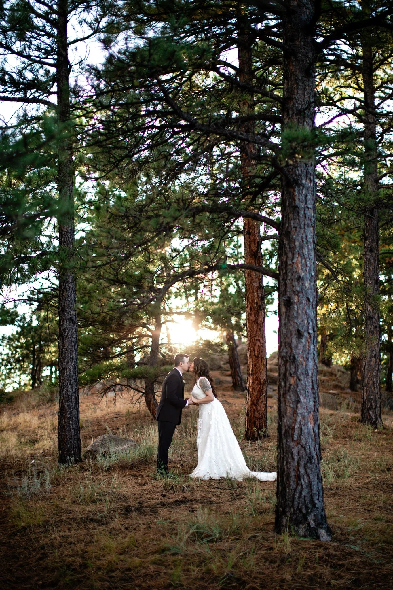 A bride and groom sharing a kiss in a forested area during sunset, surrounded by tall trees.