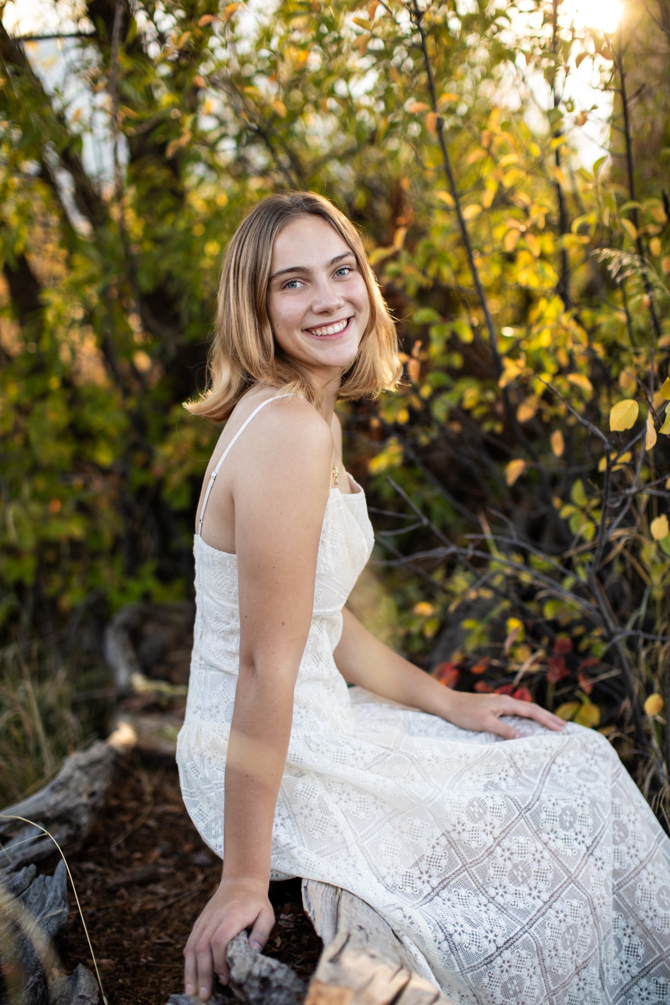 Colorado High School Senior Photographer takes a portrait of a blonde girl in a white dress sitting on a log smiling at the camera.