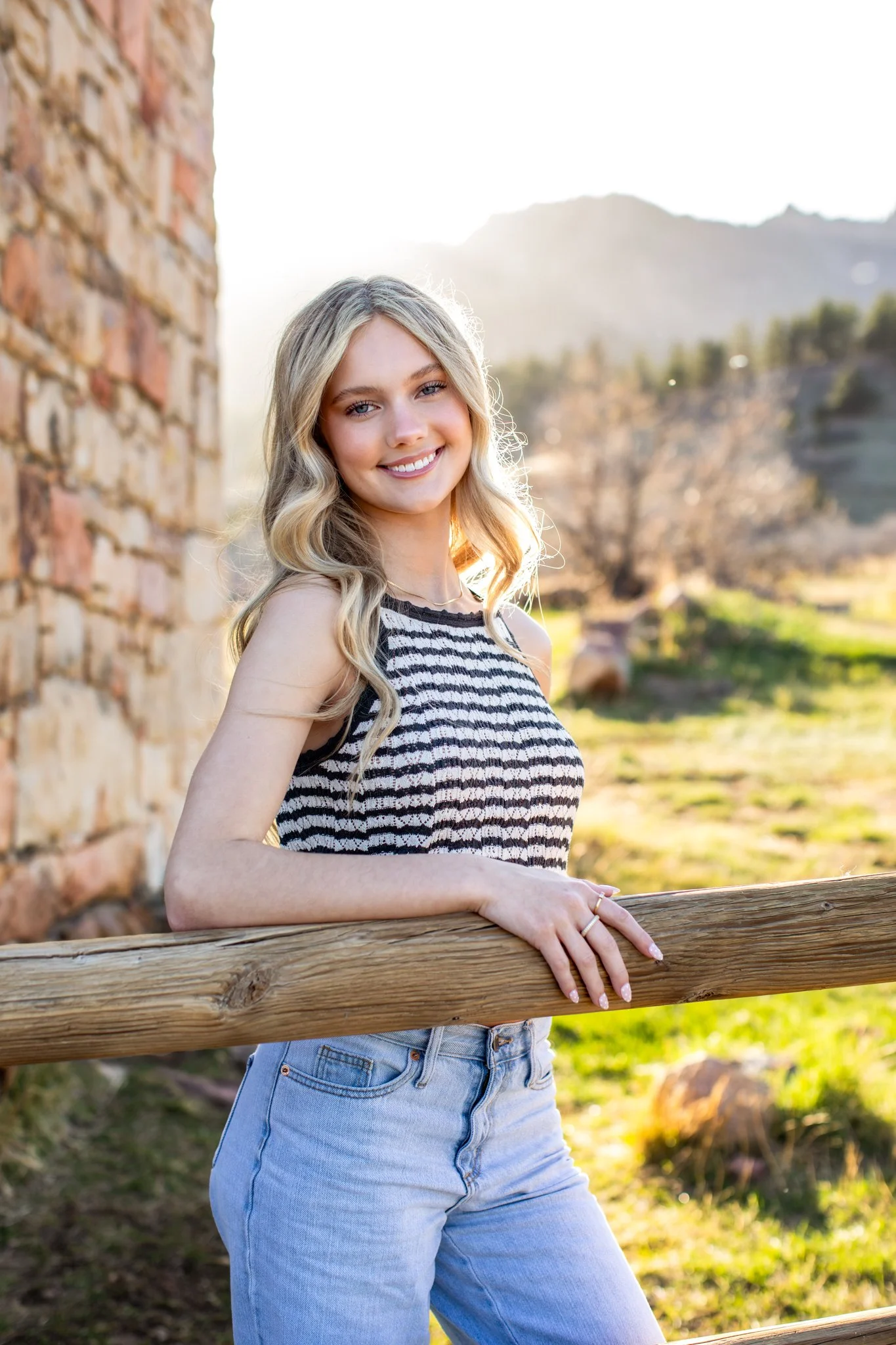 Young woman smiling outdoors near a rustic wooden fence with a stone wall, trees, and mountains in the background during sunset.