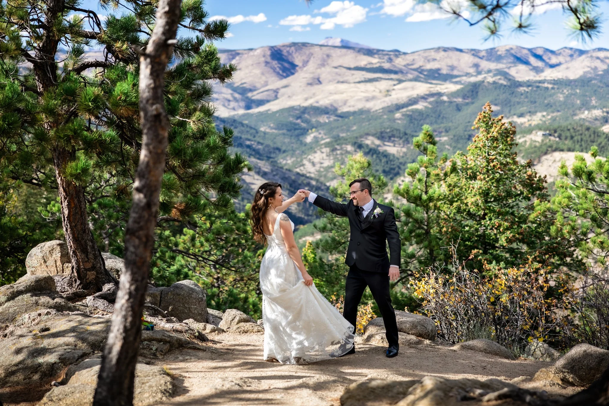 A bride in a white wedding dress and a groom in a black suit dance on a rocky trail surrounded by trees, with mountain scenery and blue sky in the background.