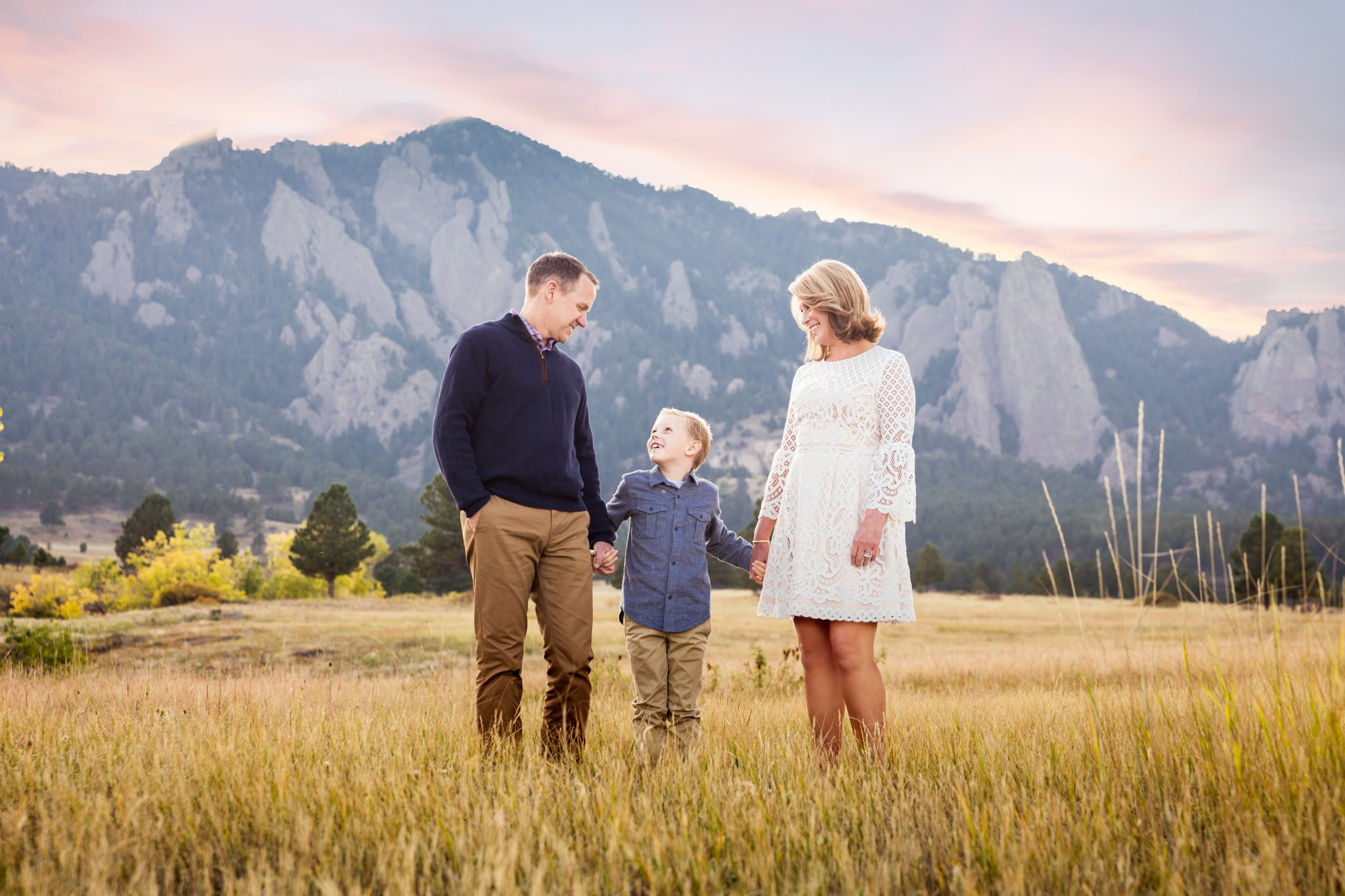 A family of three, a man, a woman, and a young boy, standing hand-in-hand in a grassy field with mountains in the background during sunset. The man wears a navy sweater and tan pants, the woman wears a white lace dress, and the boy wears a blue shirt