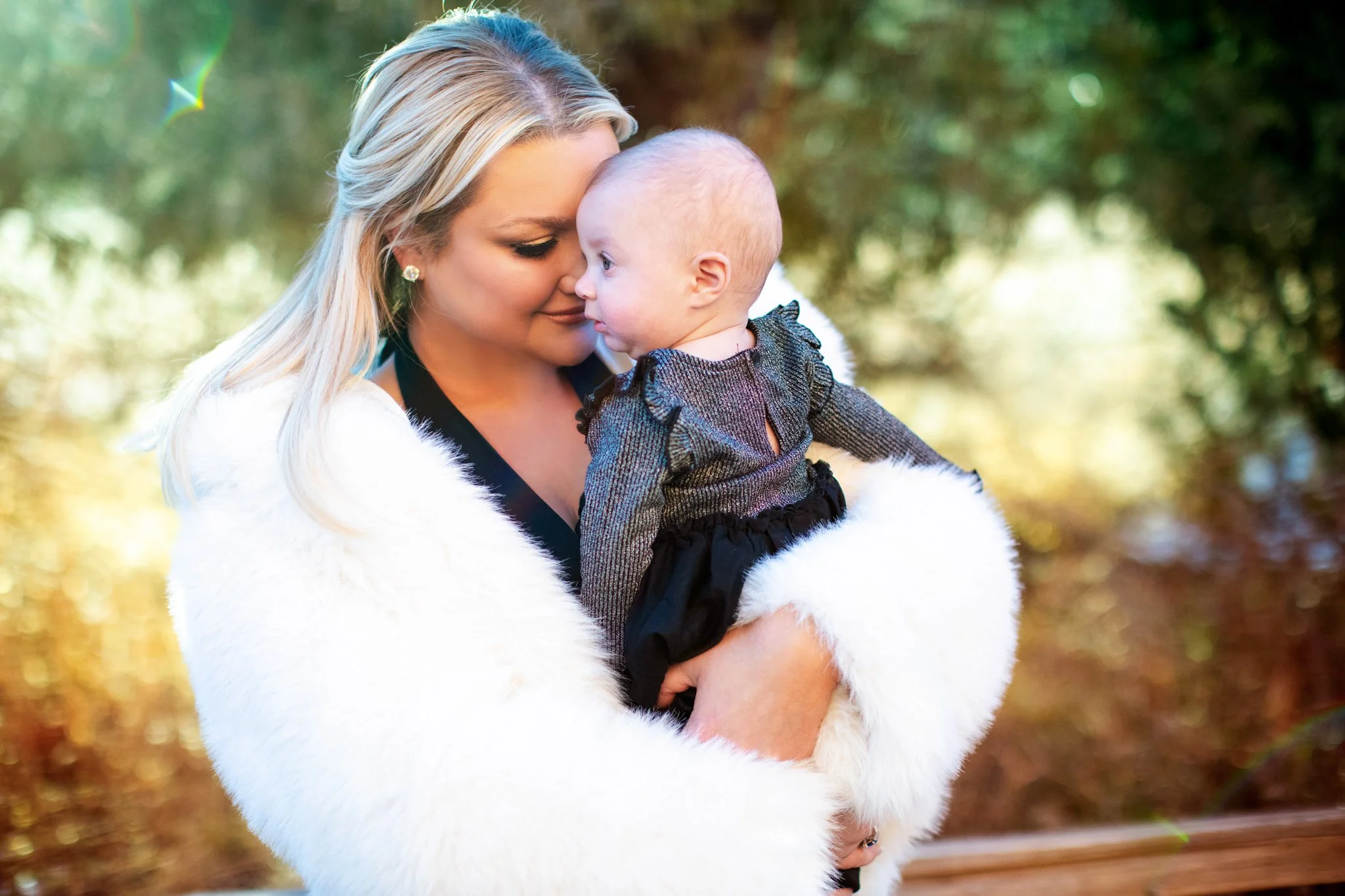 A woman with blonde hair and earrings holds a baby girl with a bald head close to her face outdoors, surrounded by trees with green leaves and sunlight.