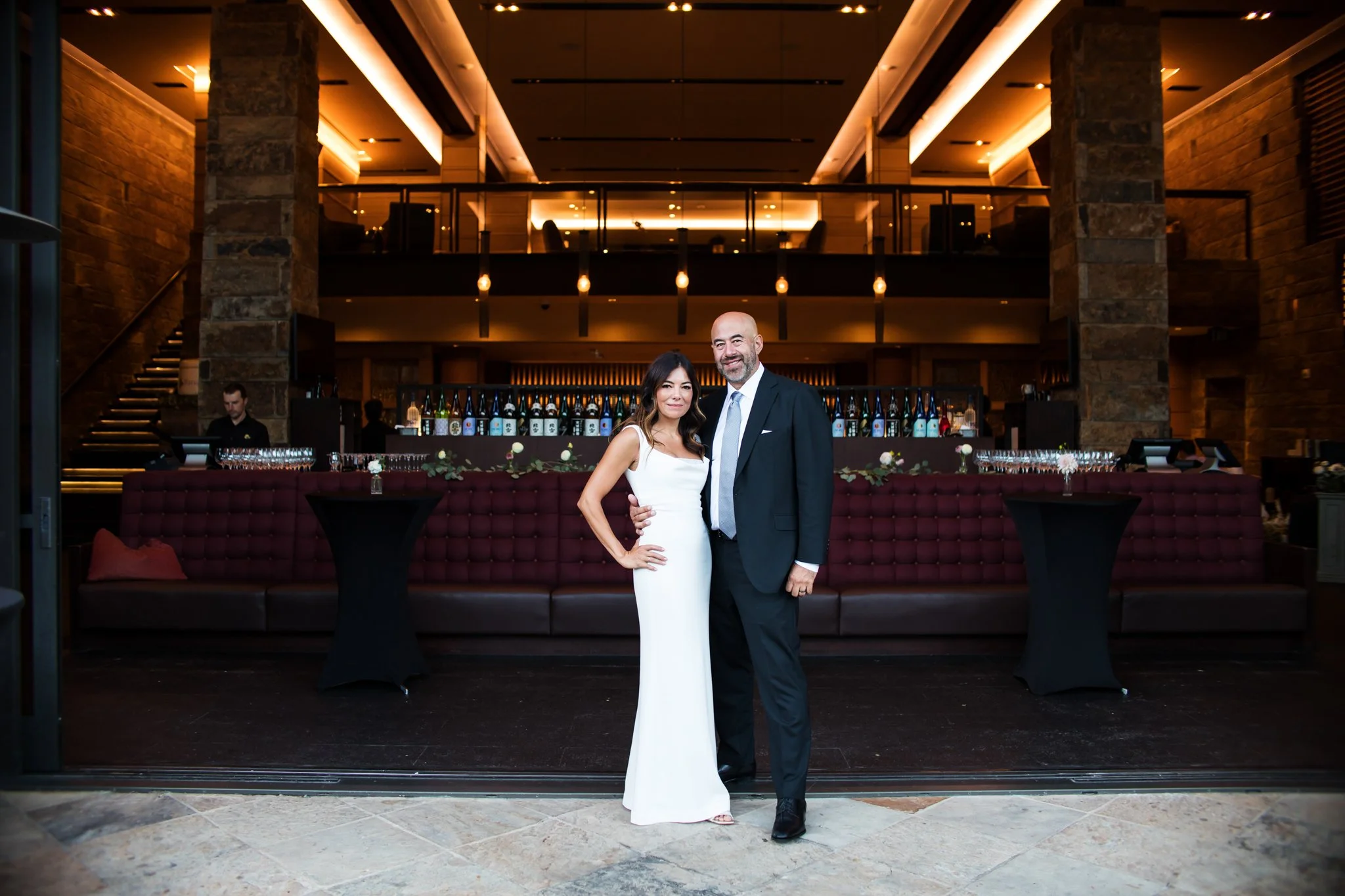 Couple dressed in formal attire standing in front of an elegant bar at a hotel or event venue, smiling at the camera.