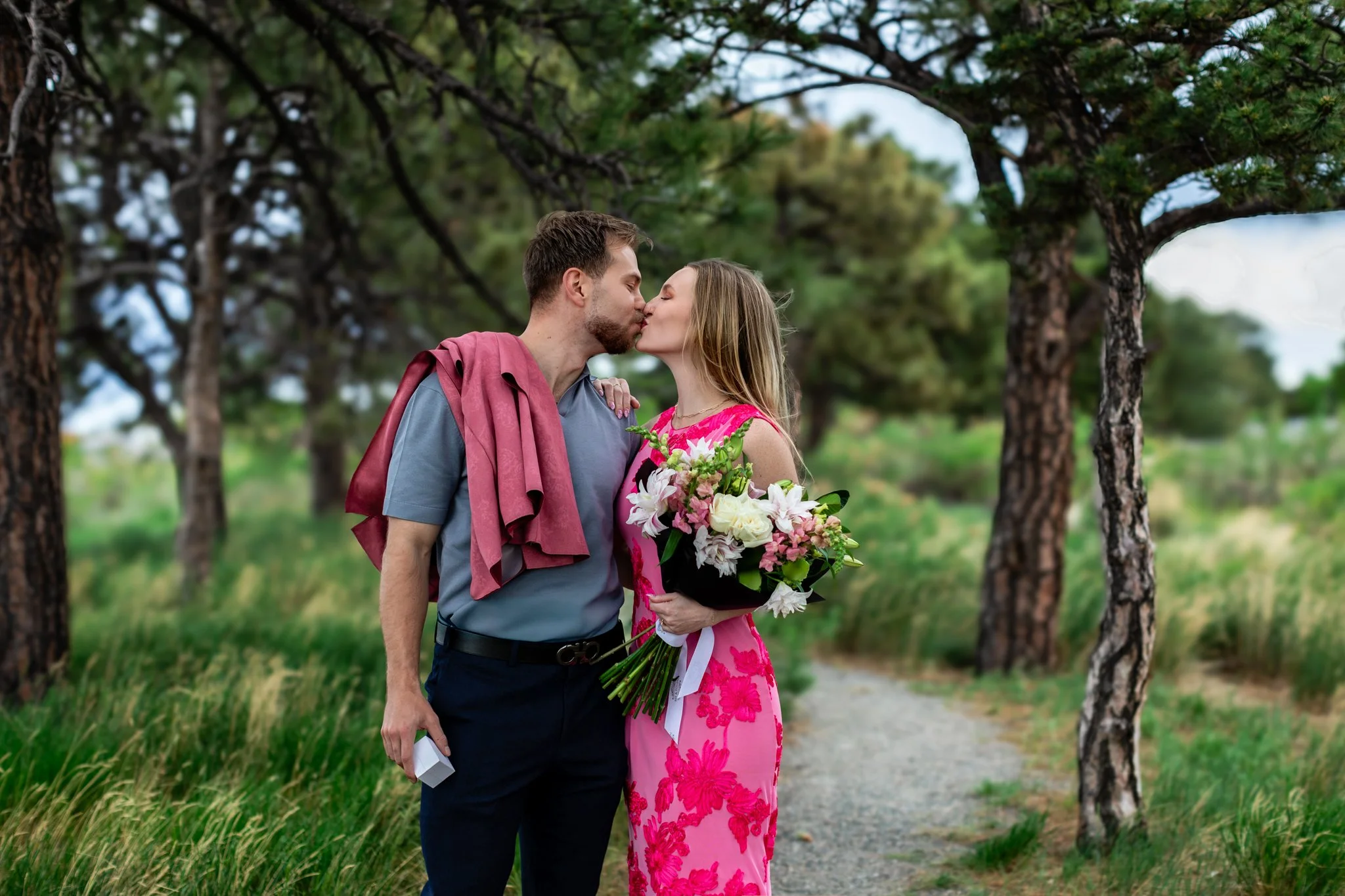 A couple sharing a kiss outdoors, with the woman holding a bouquet of flowers and the man carrying a sweater.