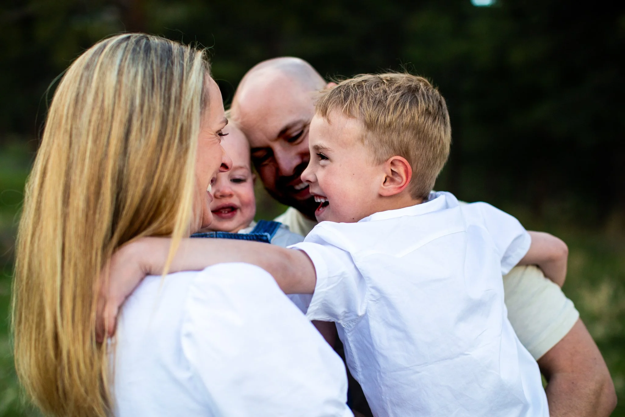 A happy family of four outdoor, embracing and smiling at each other, with blurred greenery in the background.