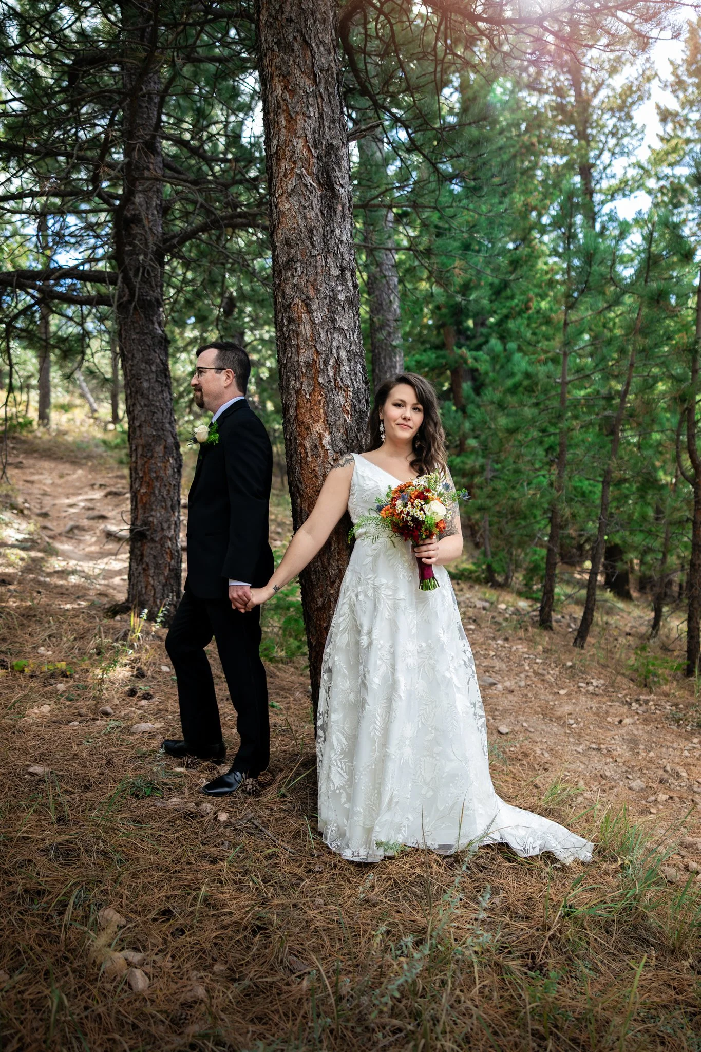 A bride and groom are standing back-to-back in a forest, holding hands. The bride wears a white wedding dress and holds a colorful bouquet, while the groom wears a black suit with a white boutonniere, looking away from the bride. They are positioned 