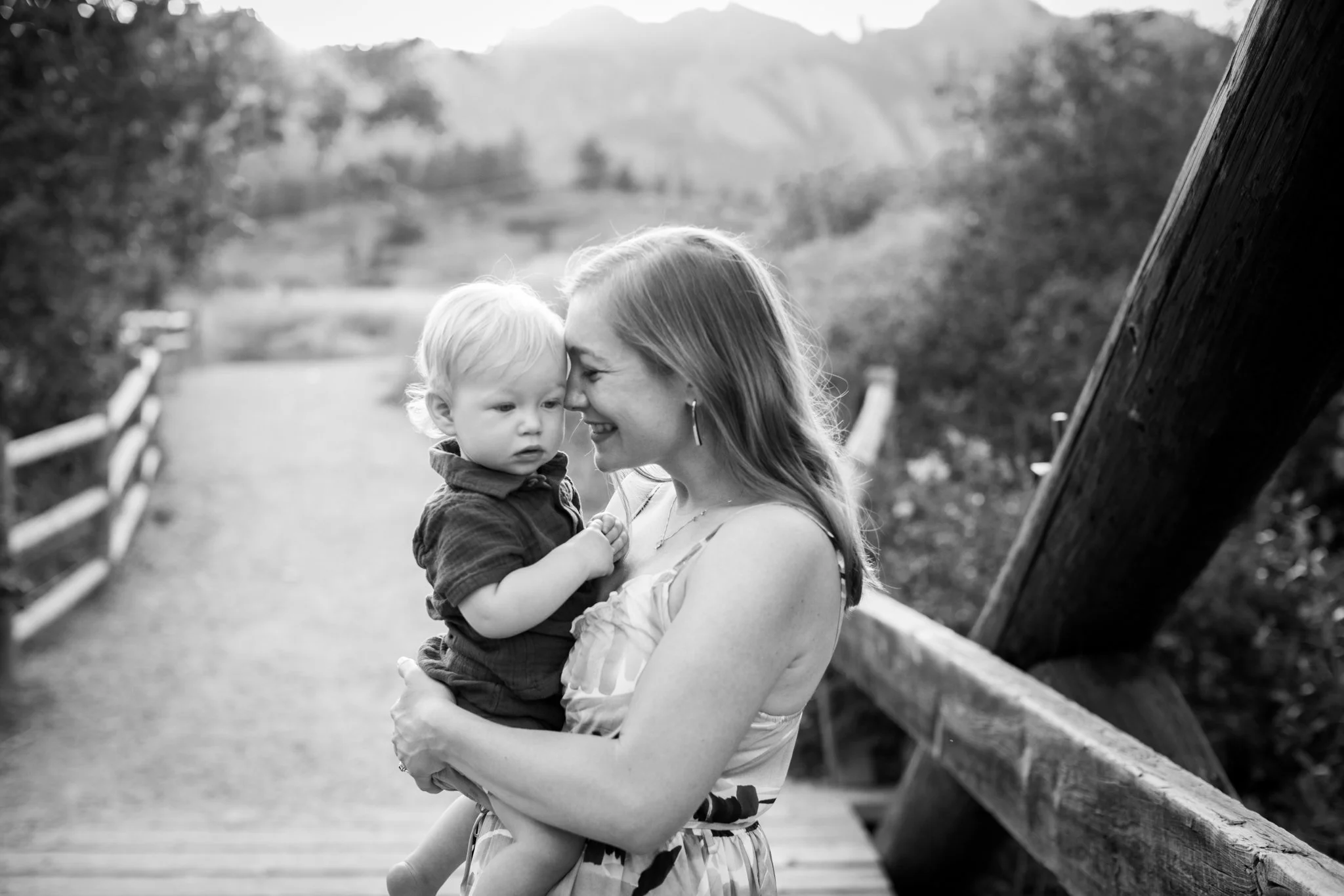 A woman holding a young child outdoors on a wooden bridge during the daytime, both smiling and touching foreheads.