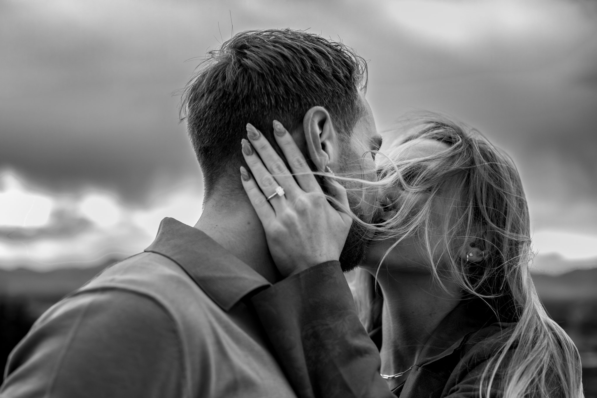 A black and white photo of a couple kissing outdoors, with the woman's hand on the man's face, showing an engagement ring, and wind blowing her hair.