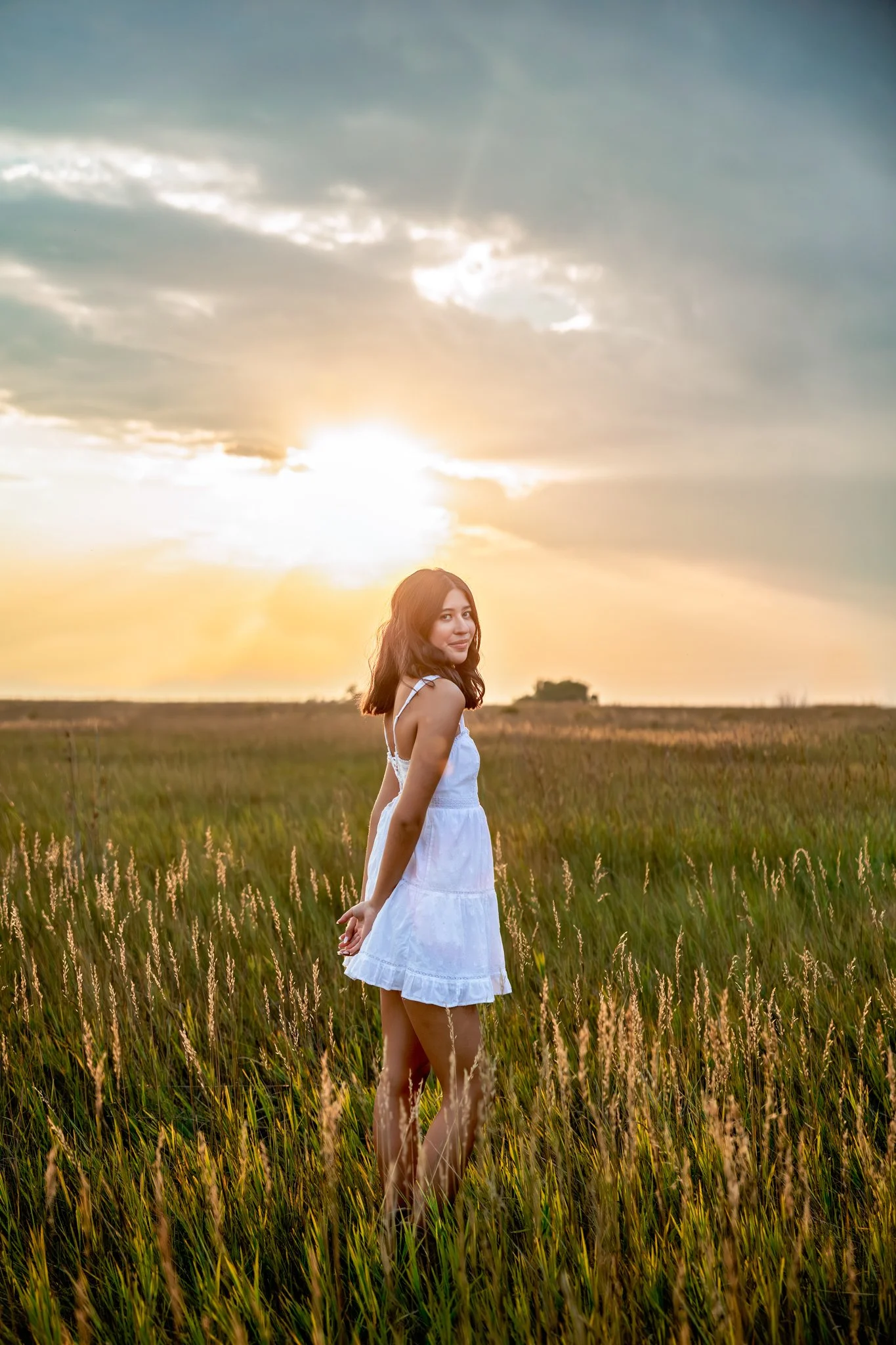 Denver High School Senior Photographer takes a portrait of high school senior girl standing in a field in a short white sundress at sunset.