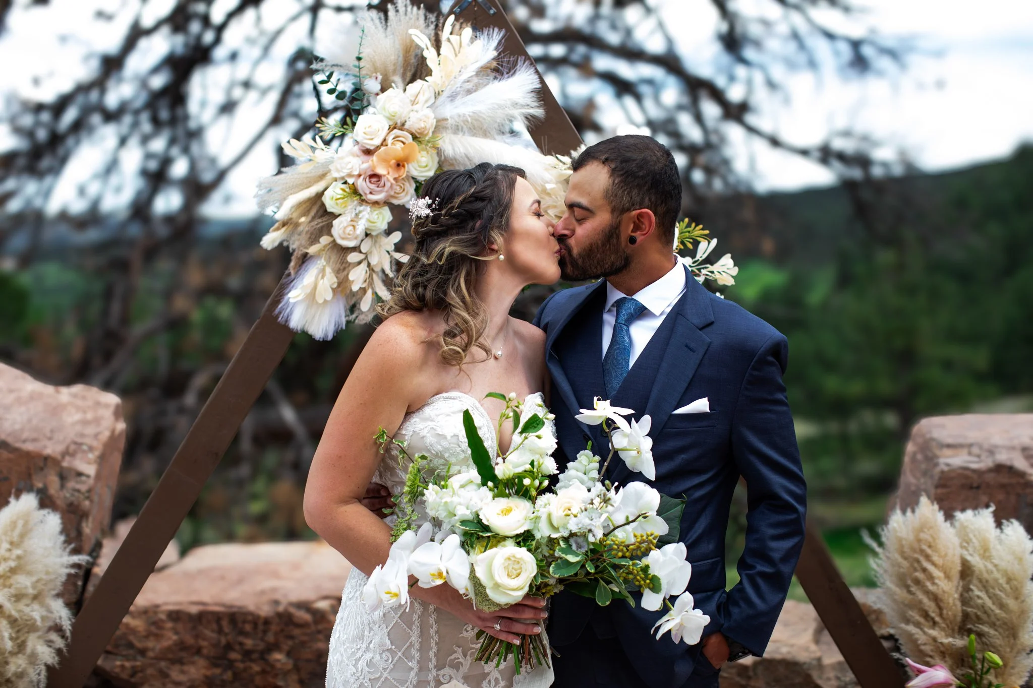 A bride and groom share a kiss during their outdoor wedding ceremony, surrounded by floral arrangements and a decorative arch with flowers and pampas grass.