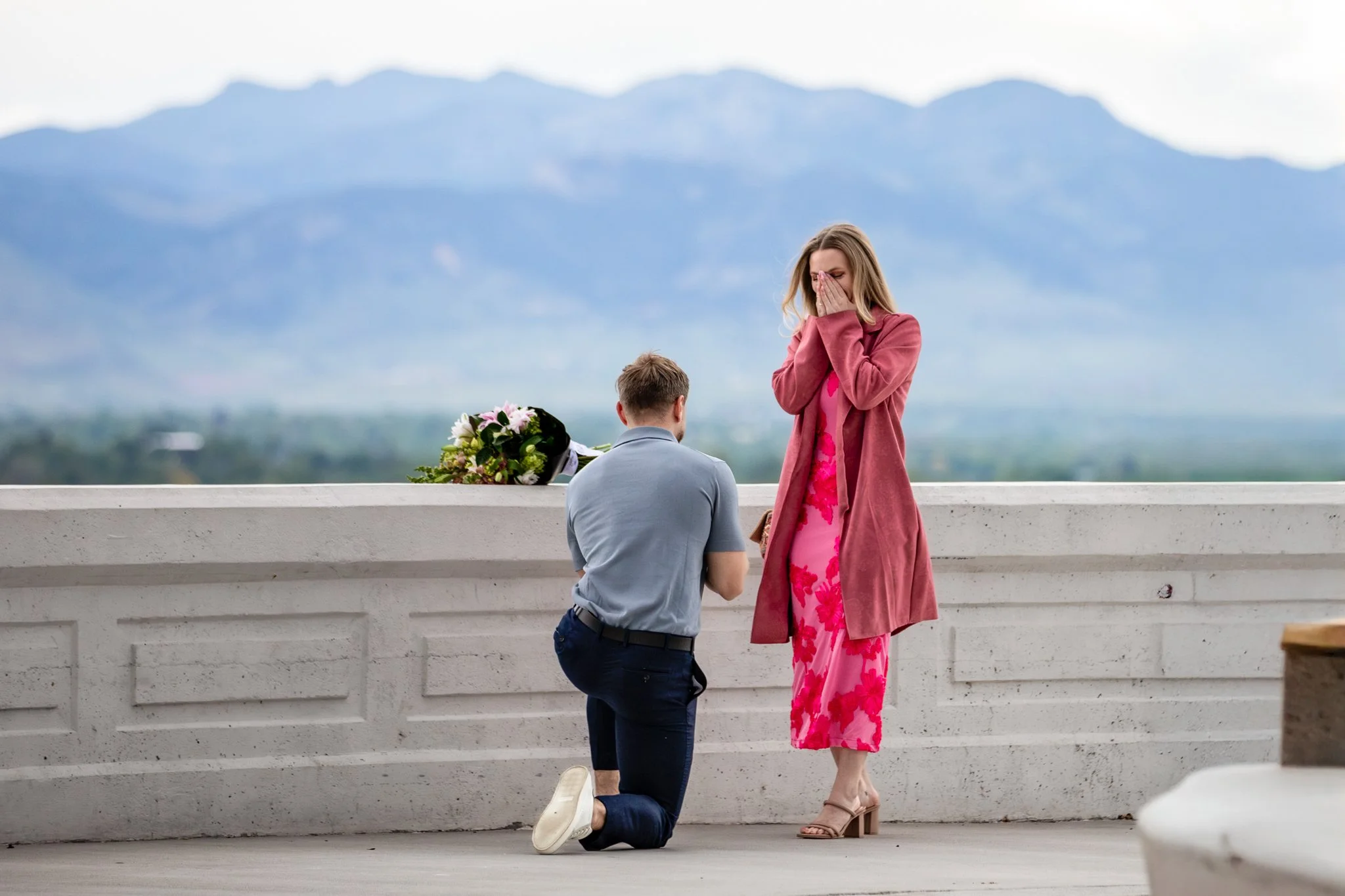 A man kneels proposing to a woman on a roadside, with mountains in the background and a bouquet of flowers on the wall nearby.