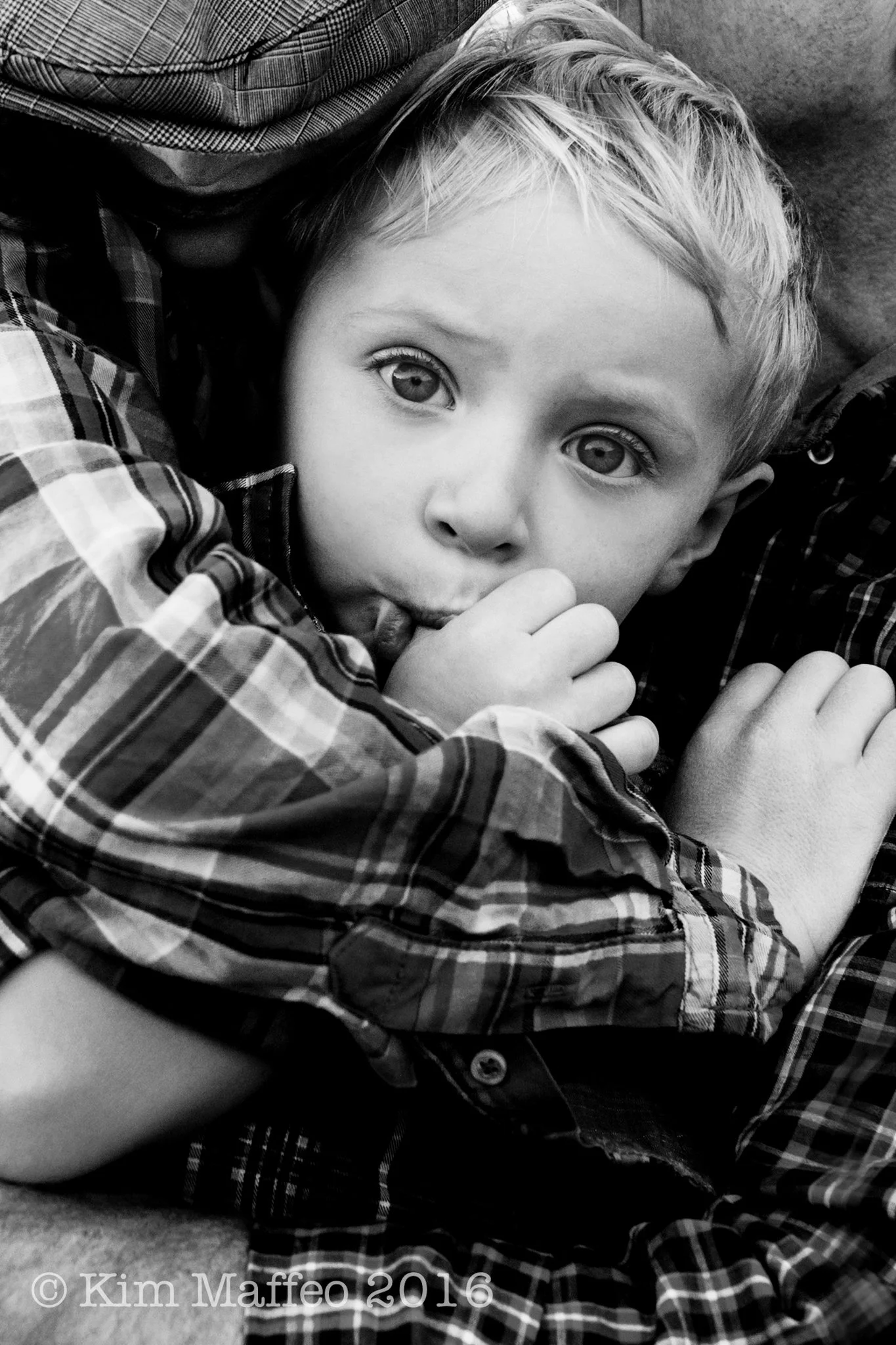 A young boy with light hair and big eyes, lying down, wearing a plaid shirt, sticking out his tongue and holding his hand near his mouth, surrounded by other people.