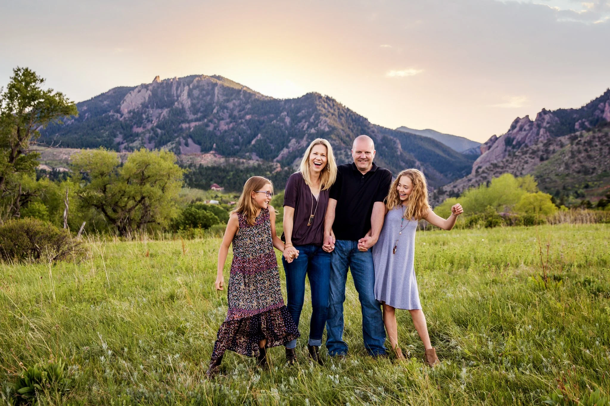 A family of four holding hands and laughing in a grassy field with mountains in the background at sunset.