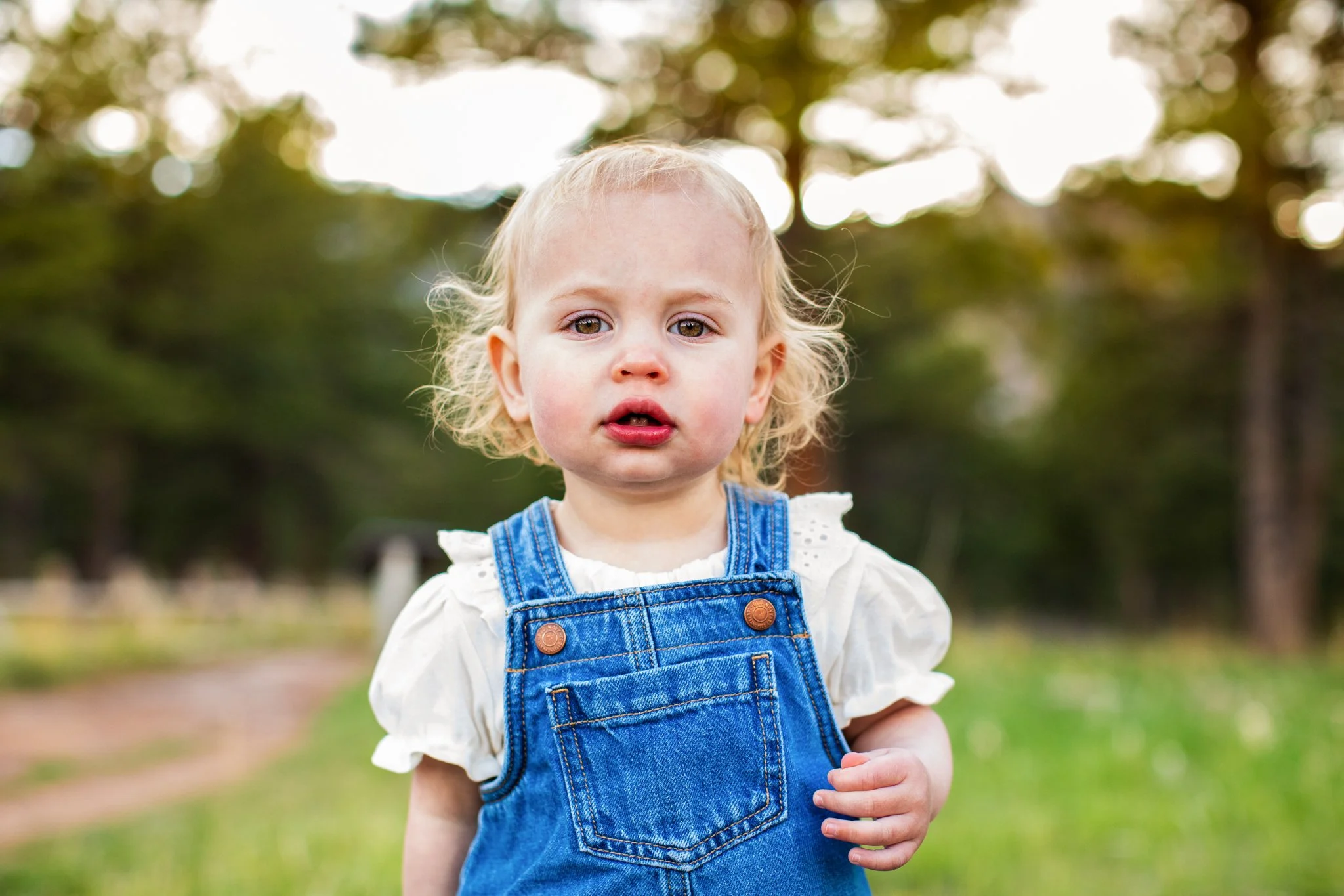 A young child with blonde curly hair wearing a white shirt and blue denim overalls standing outdoors on a grassy area with trees in the background.