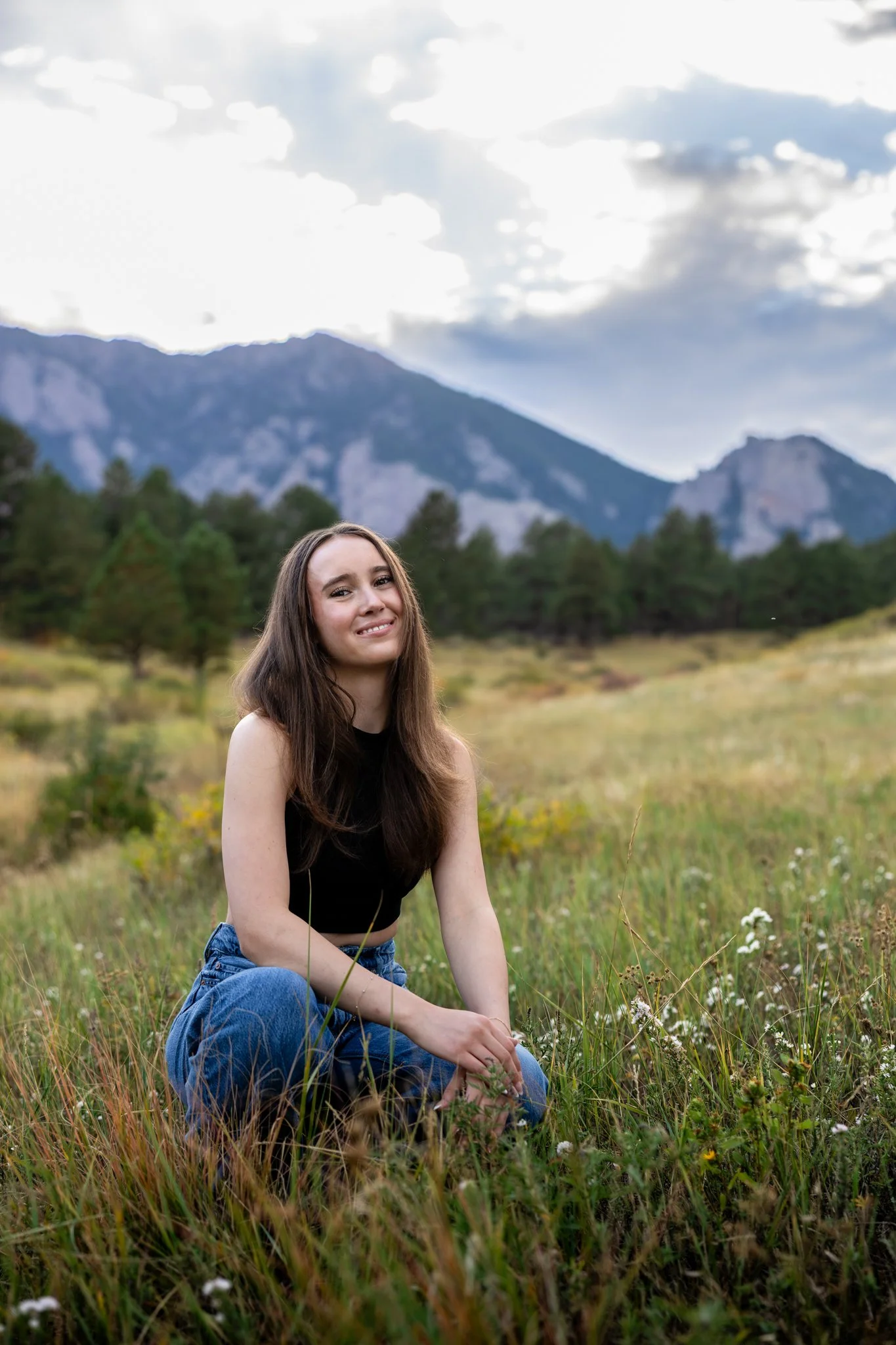 Portrait of high school girl in kneeling in a field with the Boulder flatirons in the background.