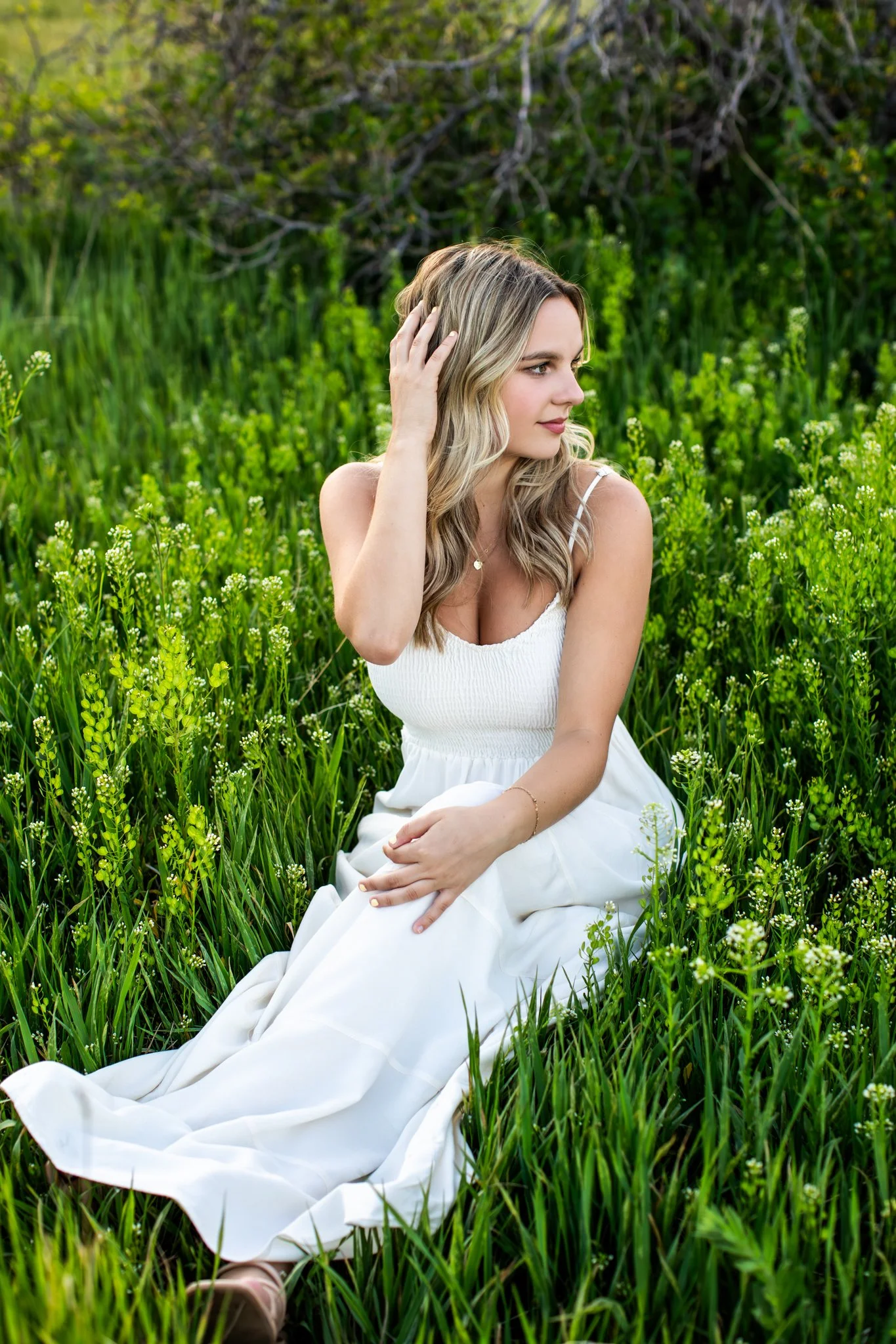 A young woman in a white dress sitting in a field of green grass and small white flowers, touching her hair and looking to the right.