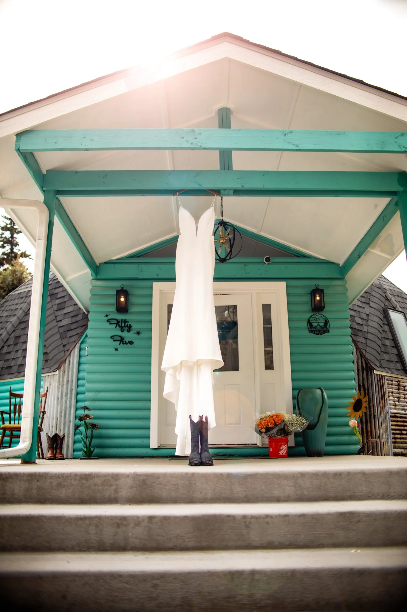 A white wedding dress hanging outside a teal-colored house, with cowboy boots placed on the steps below.