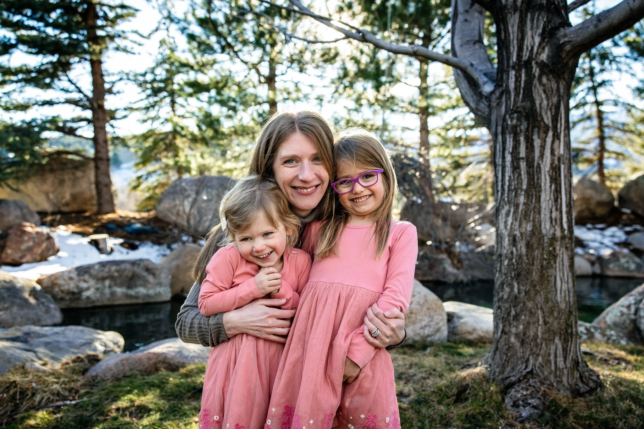 A woman and two young girls smiling and hugging outdoors next to a tree, with mountains, rocks, and a river in the background.
