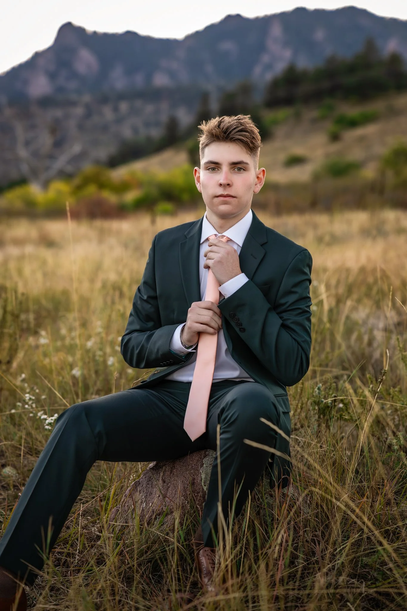 Boulder Colorado Portrait Photographer takes a picture of a young man straightening his tie in a field with the mountains behind him.