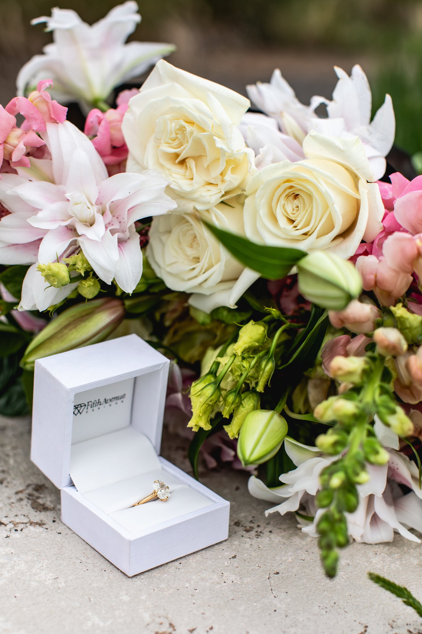 A wedding ring in a white box placed on a concrete surface with a bouquet of white, pink, and light green flowers in the background.