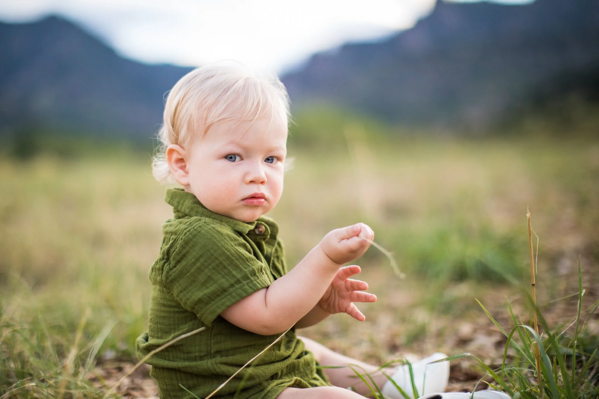 A young child with blonde hair and blue eyes sitting outdoors in a grassy field with mountains in the background, wearing a green shirt, looking at the camera.