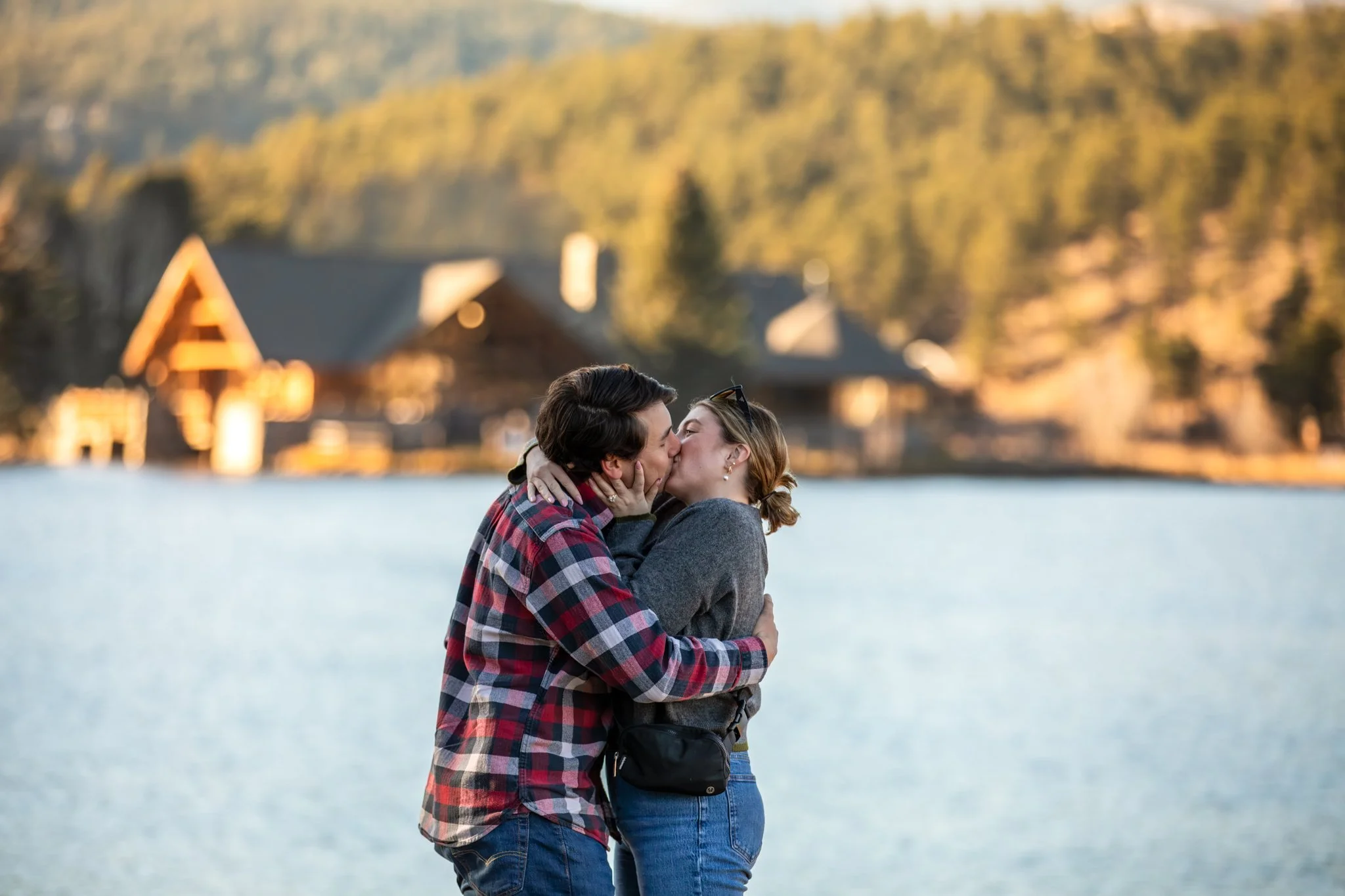 A couple kissing by a body of water with houses and trees in the background, during sunset or late afternoon.