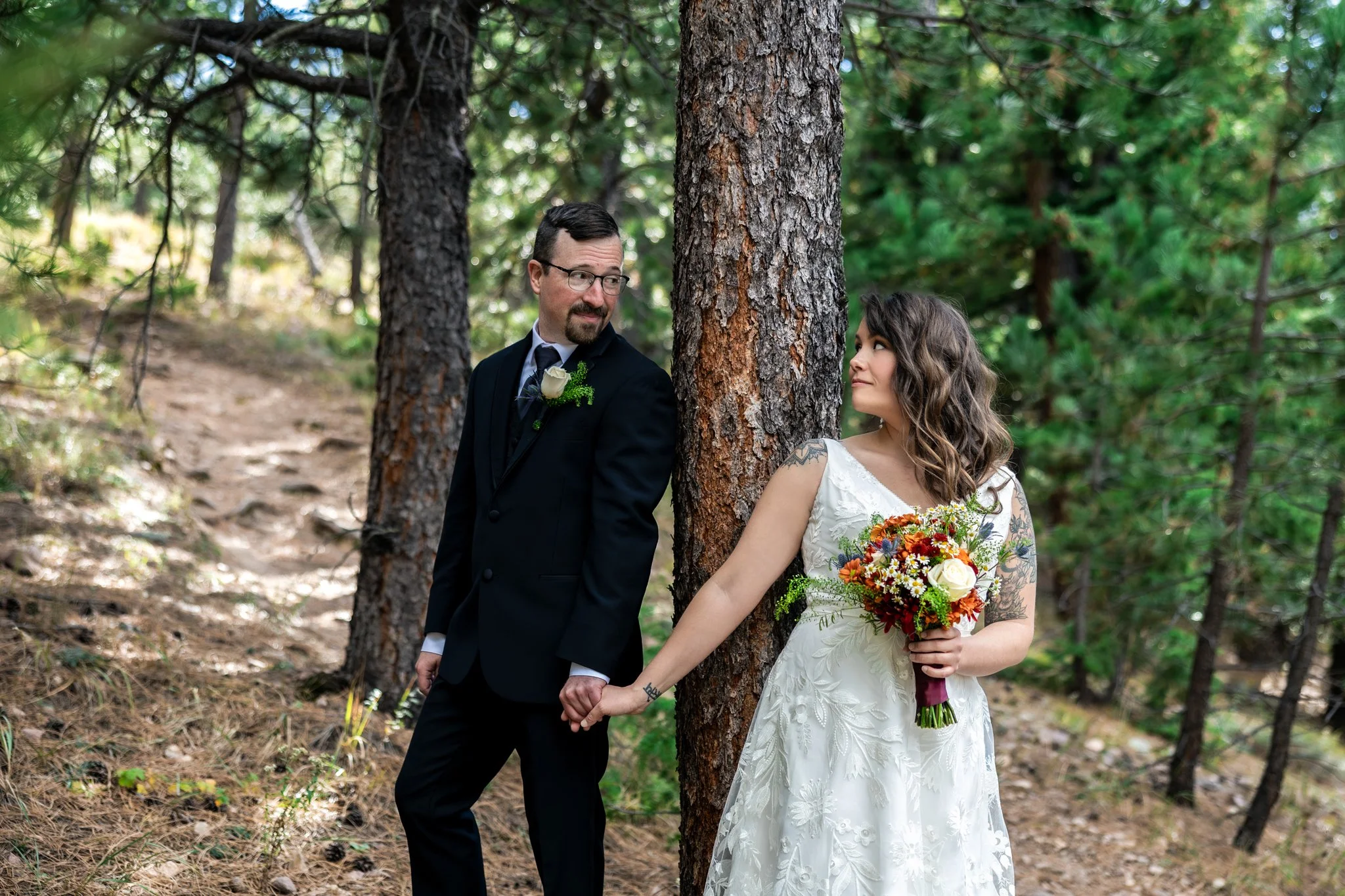 A bride and groom holding hands in a forest, standing next to a tree. The groom is wearing a black suit with a white boutonniere, and the bride is wearing a white wedding dress and holding a bouquet of colorful flowers.