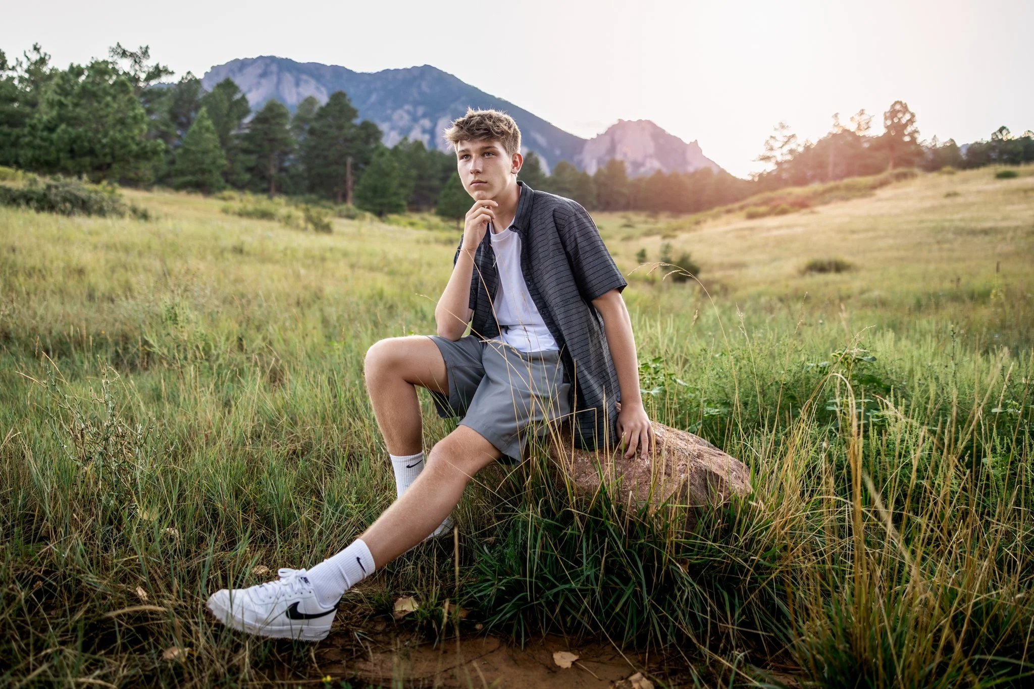 A portrait of a high school boy sitting on a rock in a grassy field with the boulder Colorado flatirons in the background.
