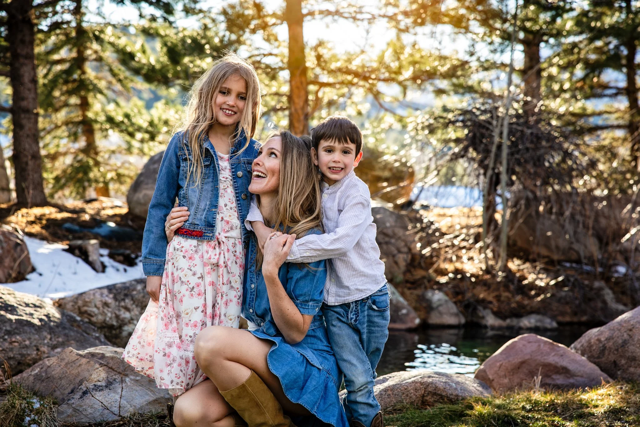A woman and two children, a girl and a boy, outdoors near a river and rocks in a forest, smiling and enjoying each other's company in sunlight.