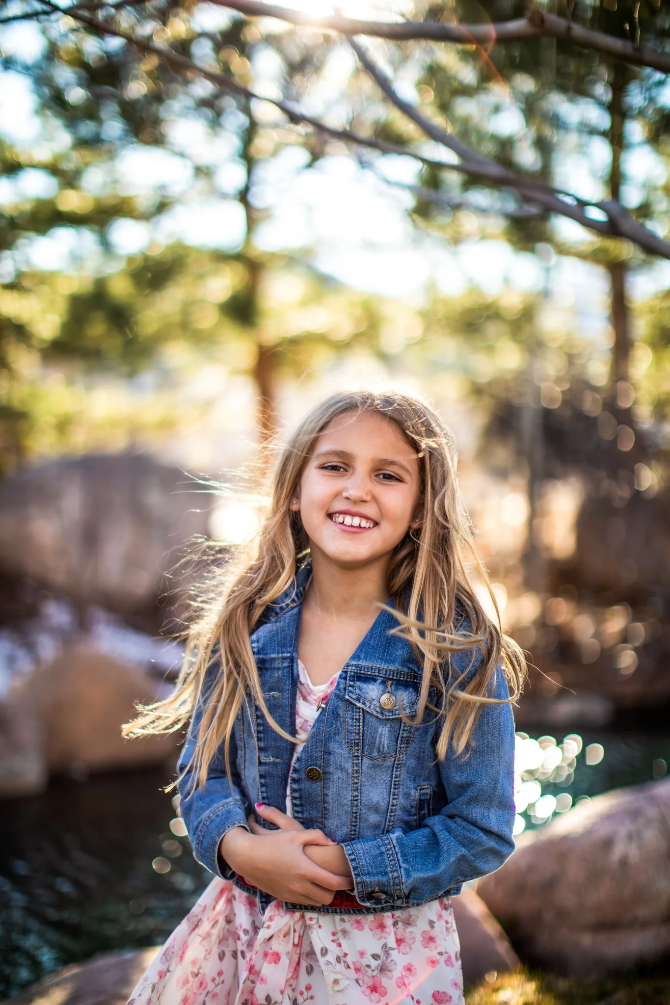 Happy young girl with long blonde hair, wearing a denim jacket and floral dress, standing outdoors near rocks and water, smiling at the camera.