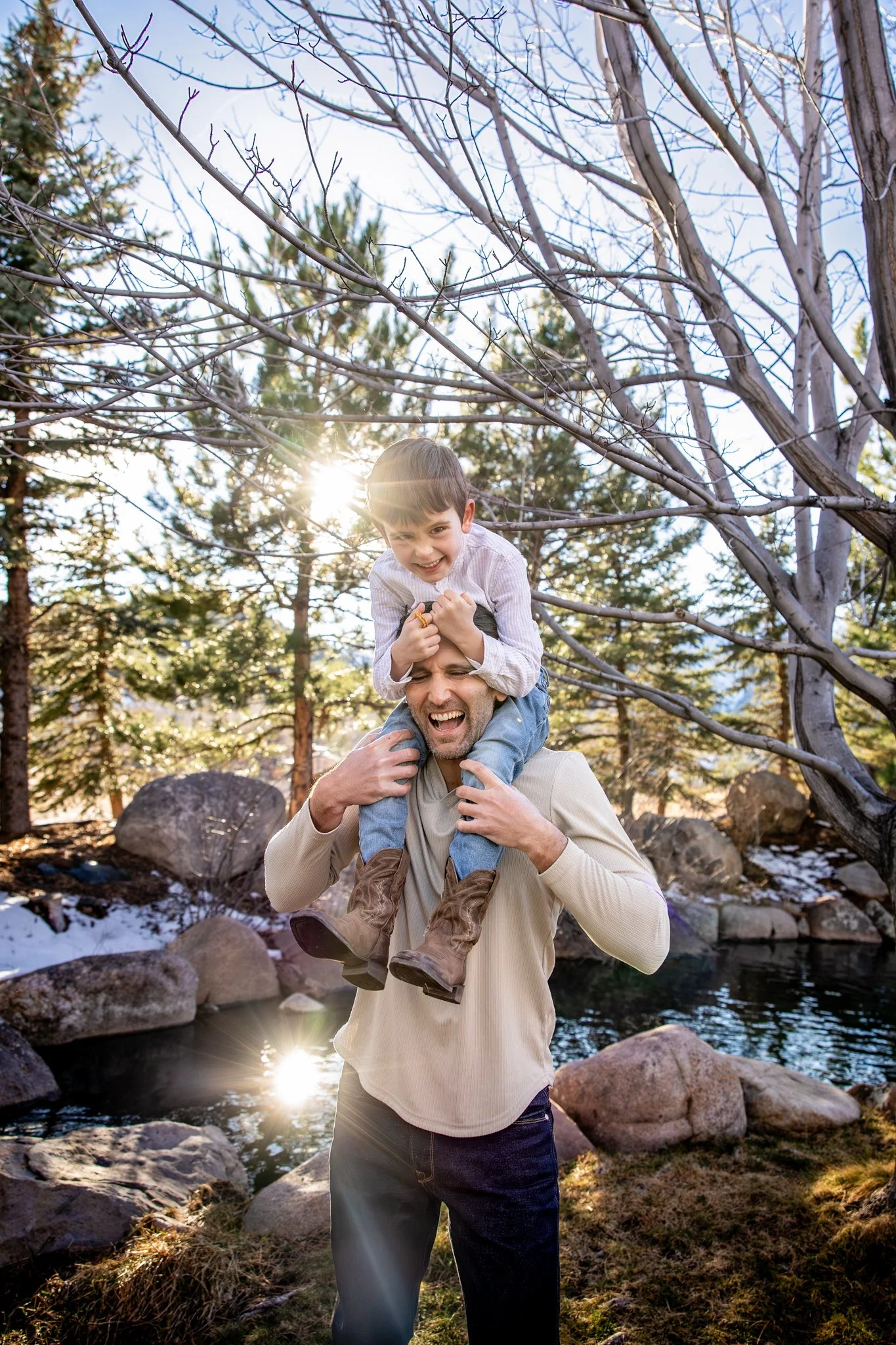 A man carrying a boy on his shoulders outdoors in a forested area with rocks and a small pond, during late afternoon sunlight.