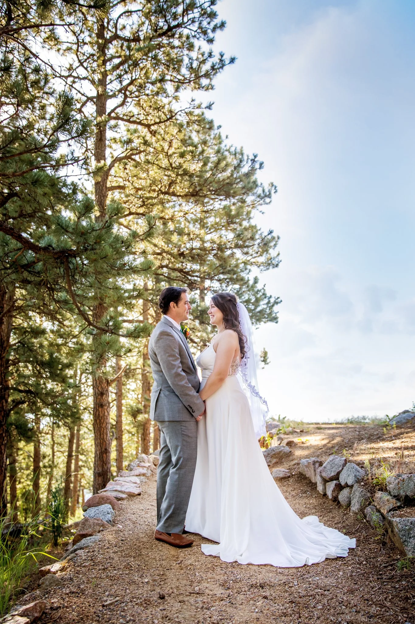 A bride and groom holding hands and smiling at each other on a dirt path in a forested area with tall trees and blue sky