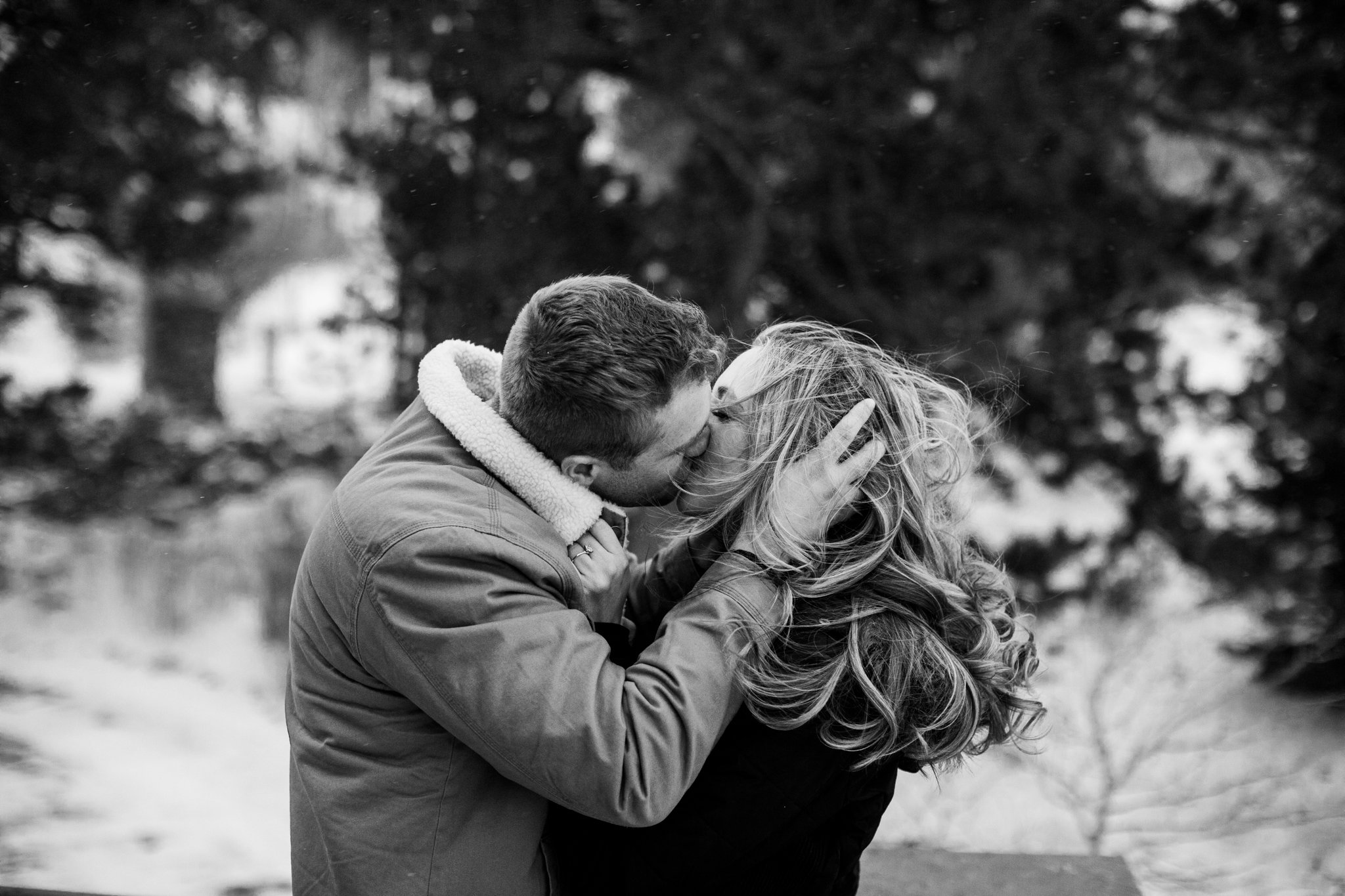 A couple kissing outdoors on a cloudy day, with the man holding the woman's face and both with closed eyes, surrounded by blurred trees.