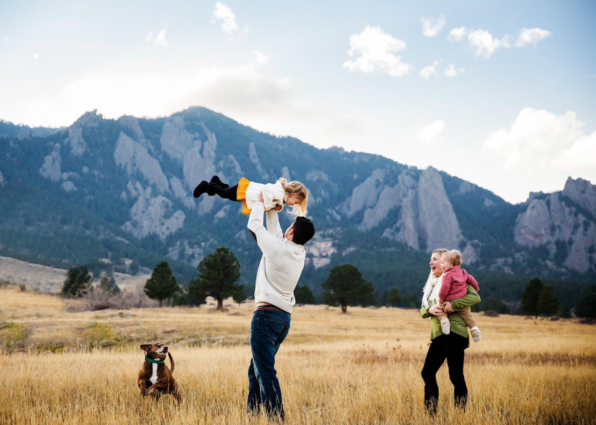 Family of four with two children and a dog enjoying a day outdoors in a grassy field with mountains in the background. The father is lifting a young girl, and the mother is holding a younger child, all smiling.