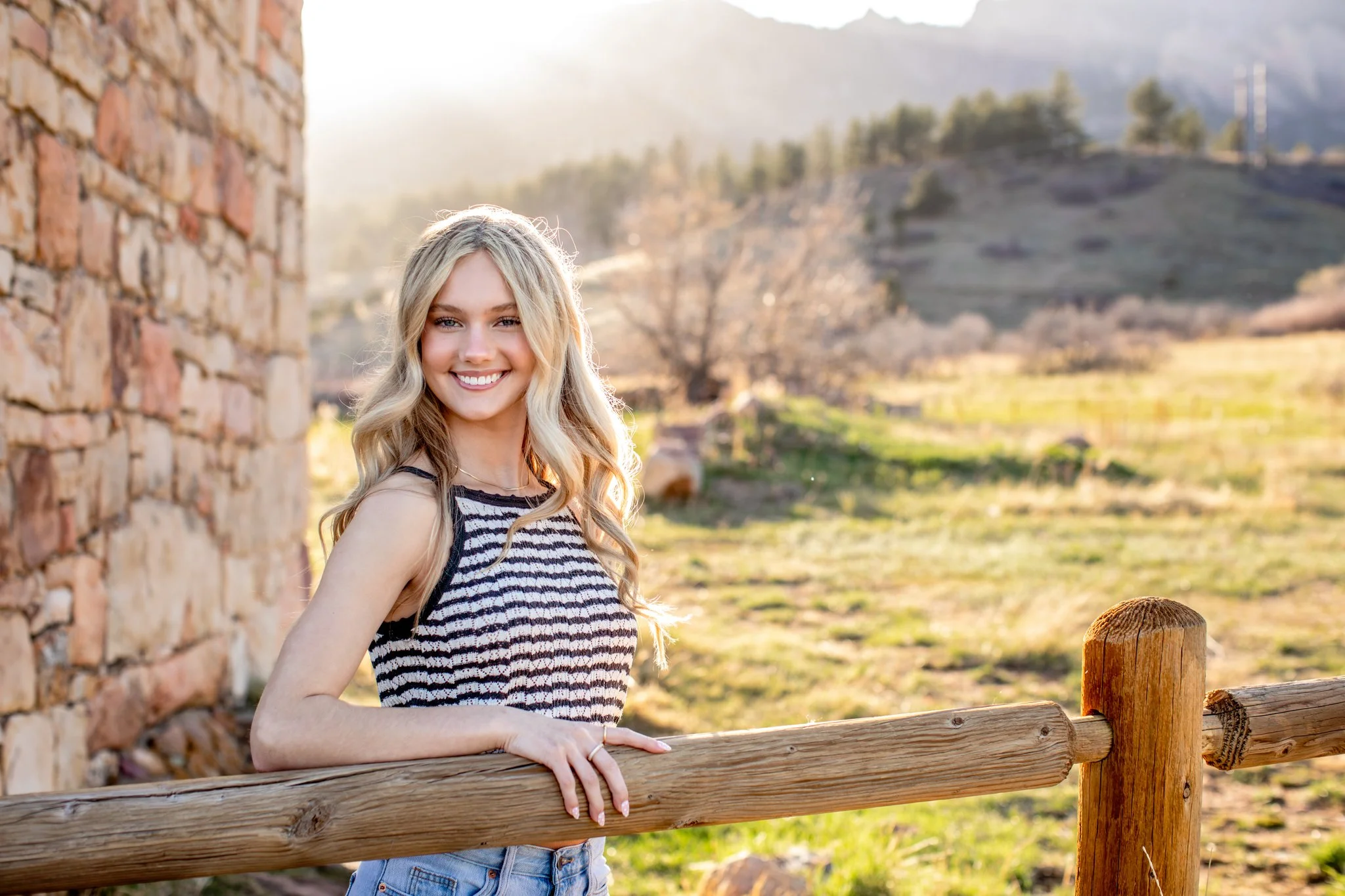 Colorado portrait photographer captures a picture of a blonde girl wearing a black and white striped tank top leaning against a fence at sunset.