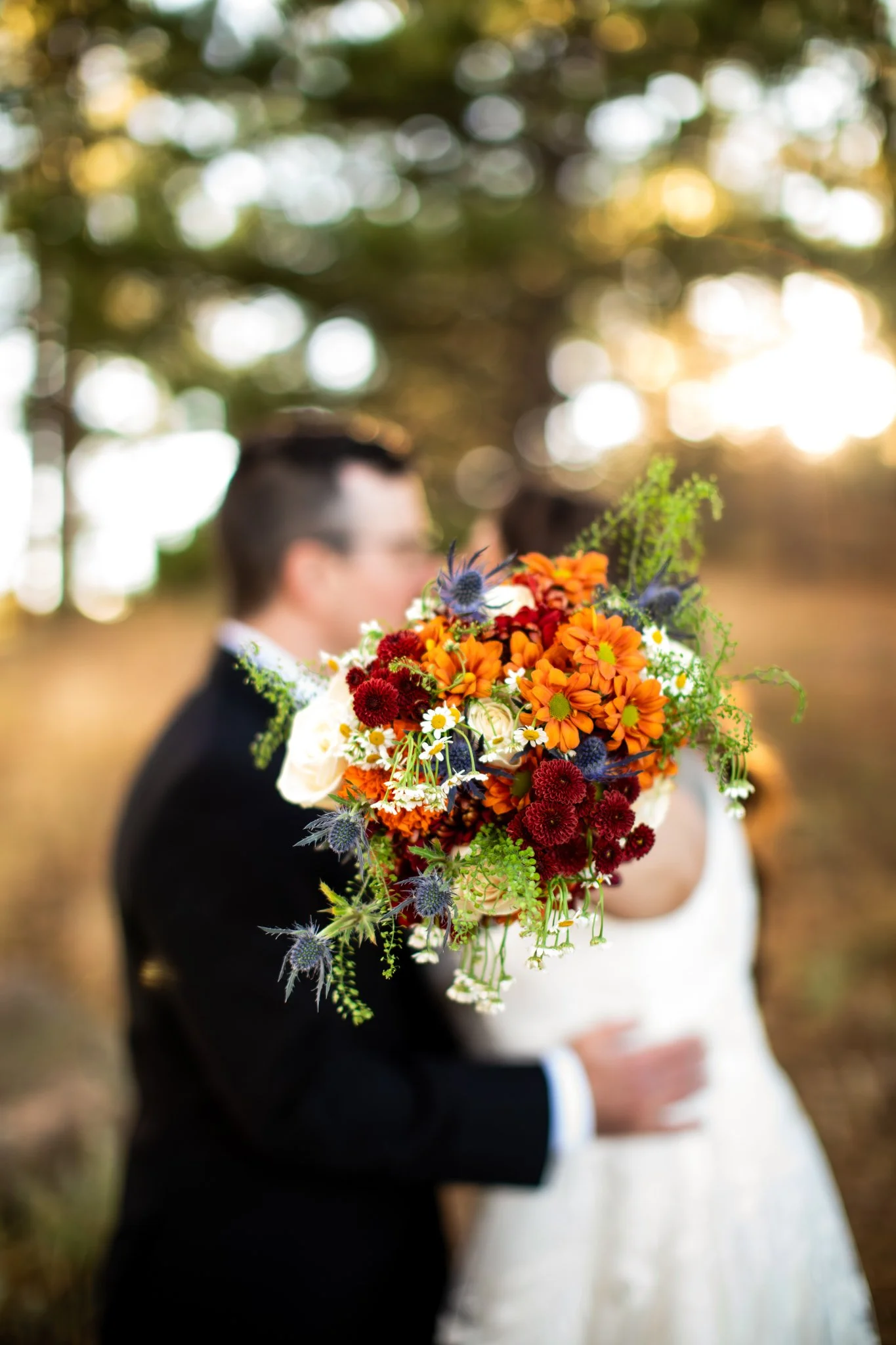 A floral bouquet with orange, red, white roses, and various greens, held in front of a blurred couple in wedding attire outdoors at sunset.
