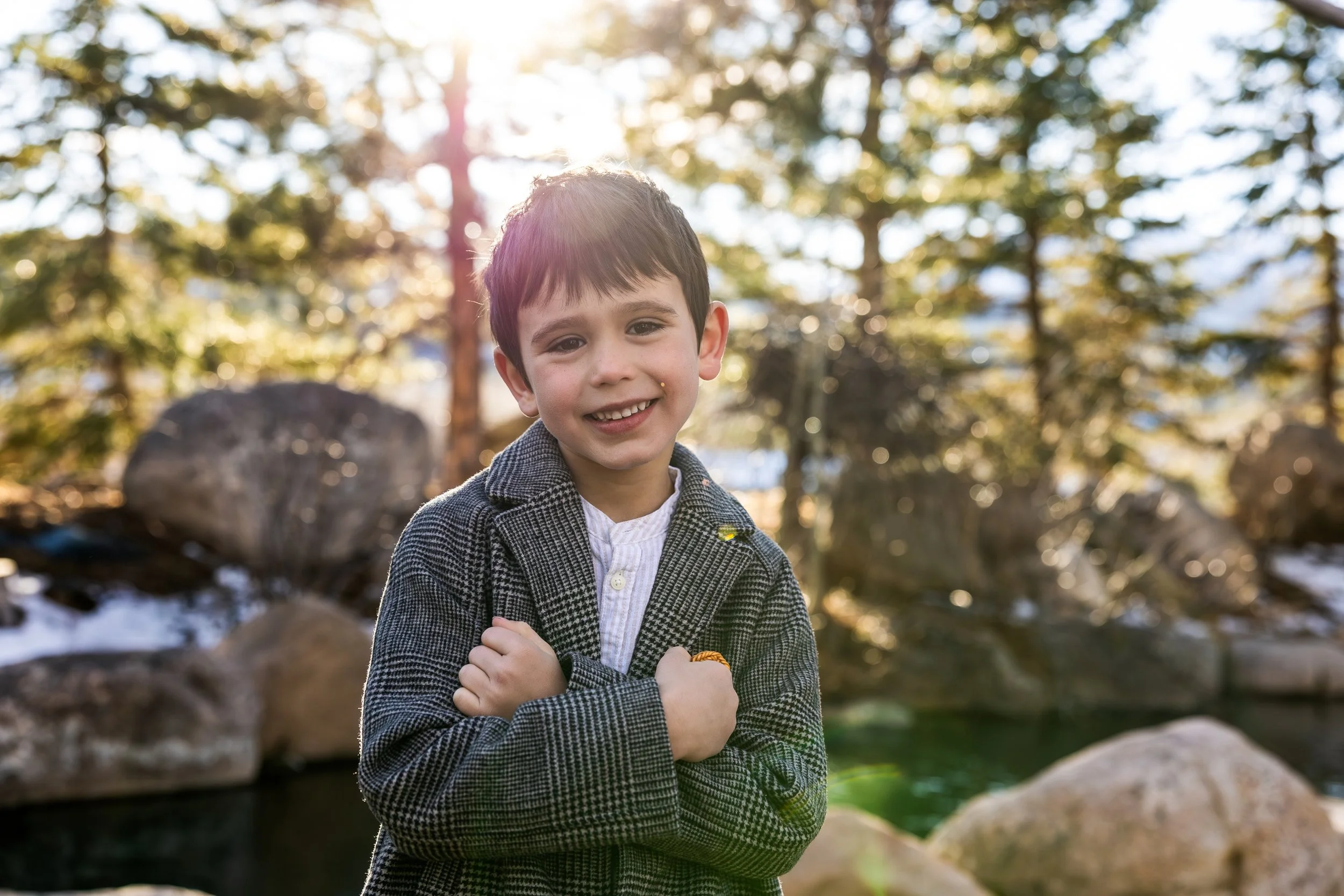 A young boy outdoors with sunlight filtering through trees in the background, smiling at the camera with arms crossed.
