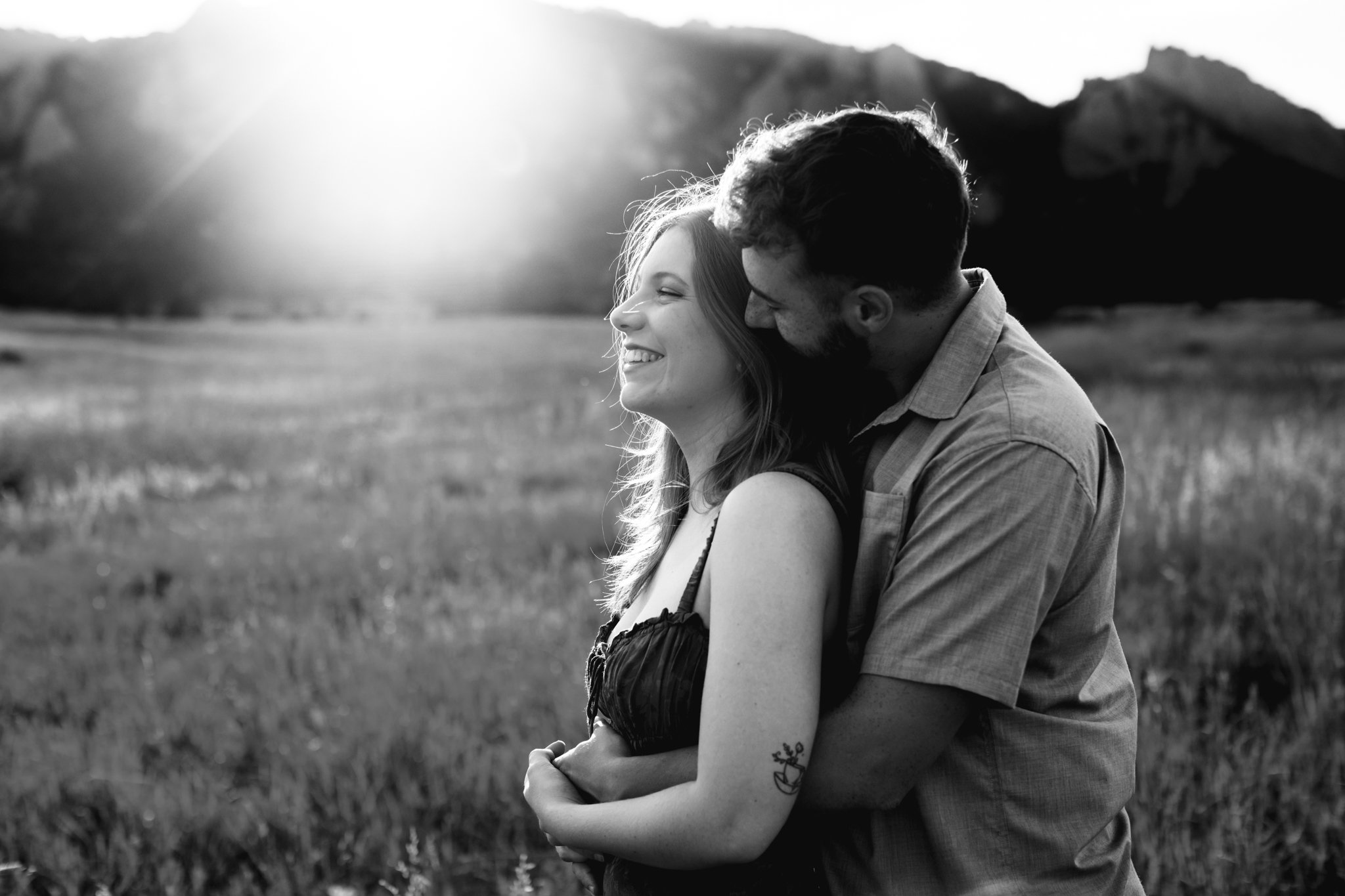 A black and white photo of a smiling woman and a man hugging her from behind in an open field with mountains in the background at sunset.