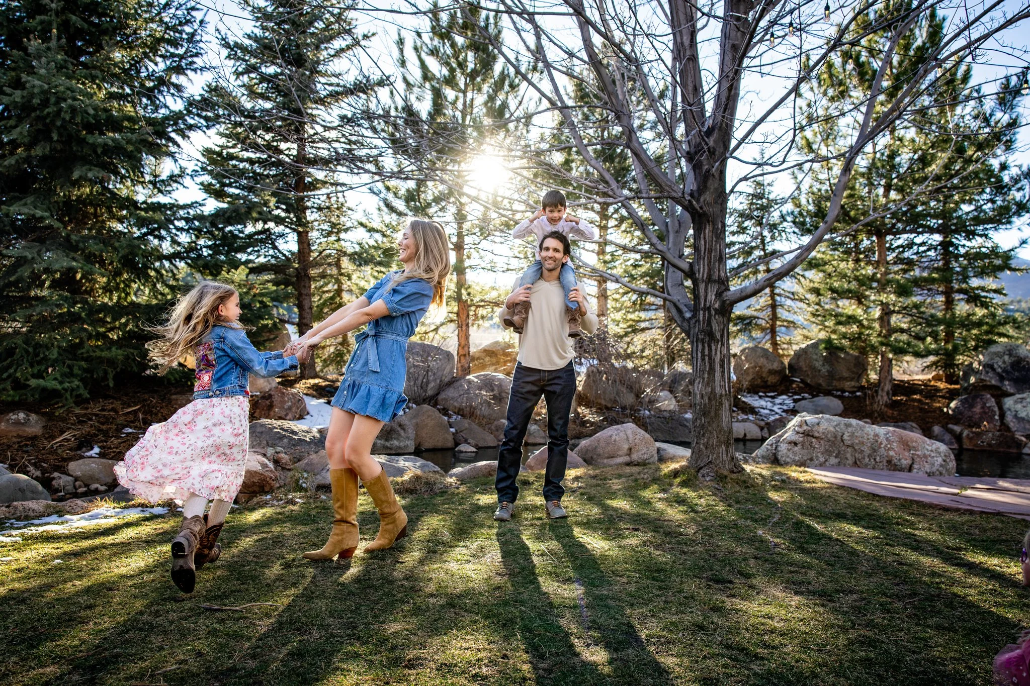 Family of four outdoors enjoying playtime on a sunny day with trees in the background, a woman and girl are dancing and holding hands, a man holds a boy on his shoulders near a leafless tree.