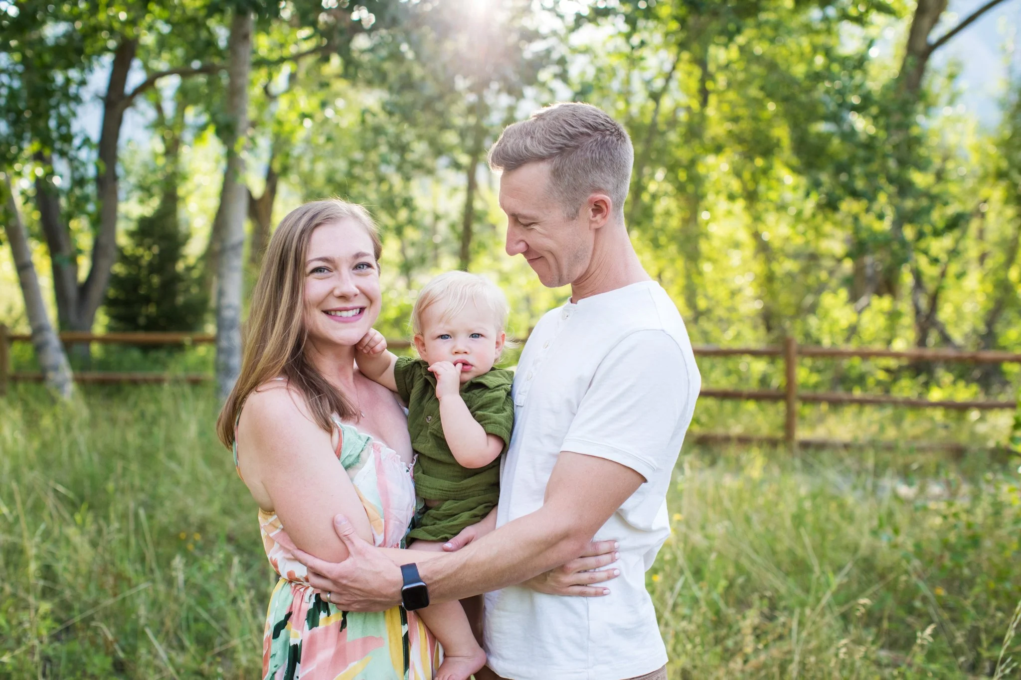 A family of three outdoors in a grassy area with trees and sunlight, with the mother holding a young boy, and the father standing close to them, all smiling.